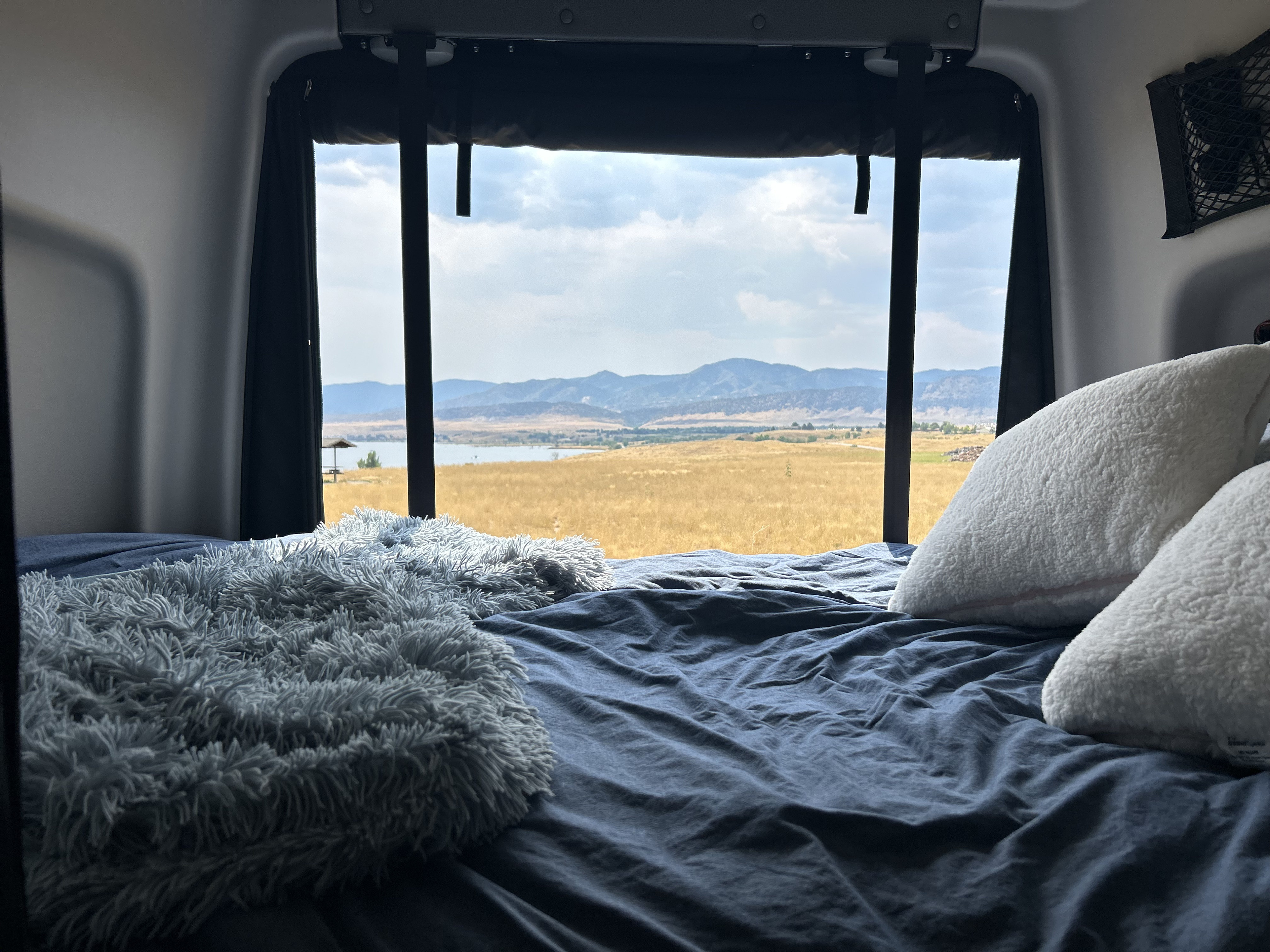 Cozy RV rental bed with a panoramic view of a lake, golden fields, and mountains under a cloudy sky.. Winnebago Revel 2023