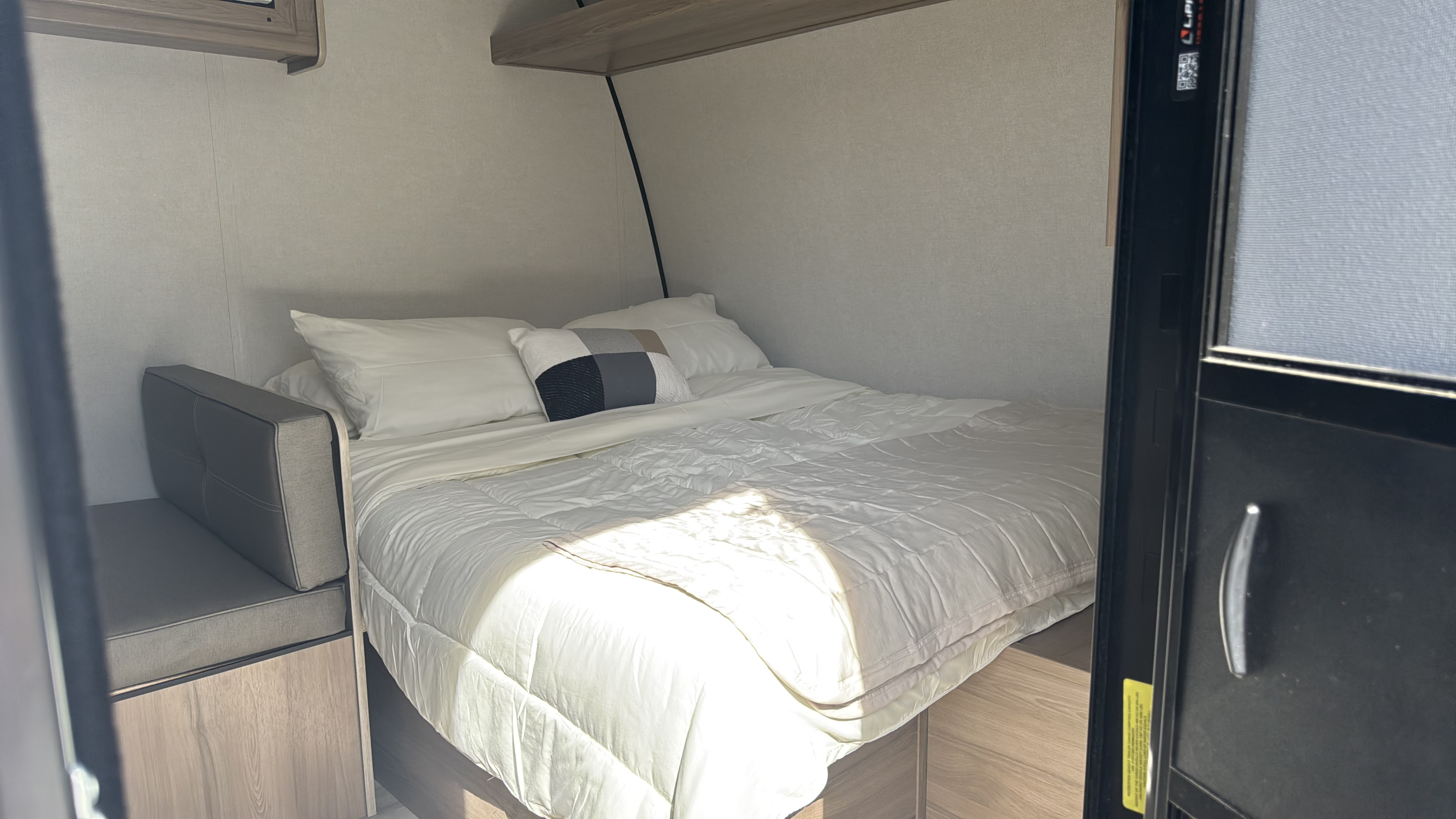 RV rental bedroom with clean white bedding, pillows, and a modern side bench. Sunlight illuminates the cozy interior.. Keystone Coleman 2025