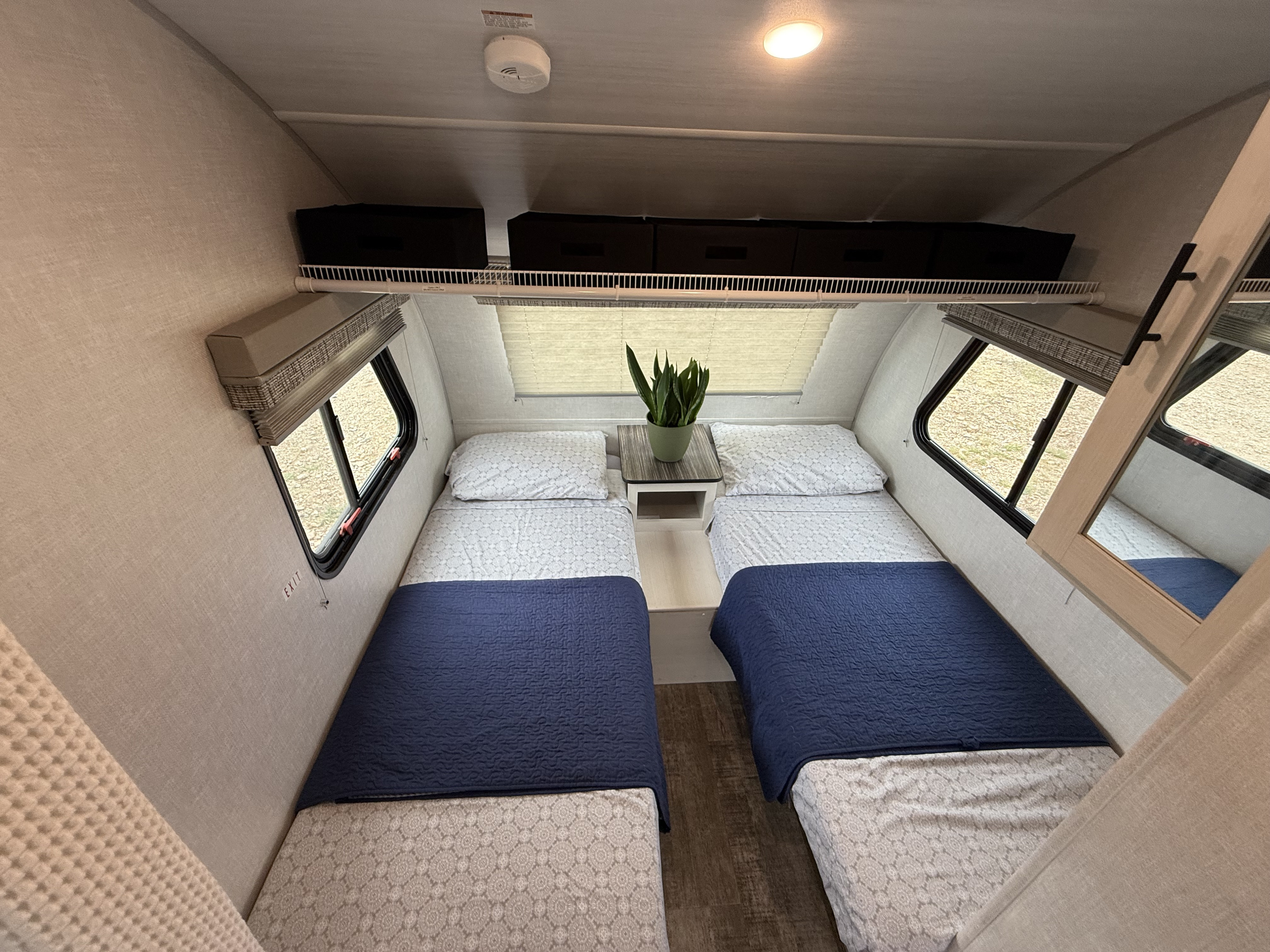 An RV rental bedroom shows two single beds with blue quilts, a nightstand, windows, and an overhead shelf with storage bins.. R-Pod R-Pod Trailer 2021