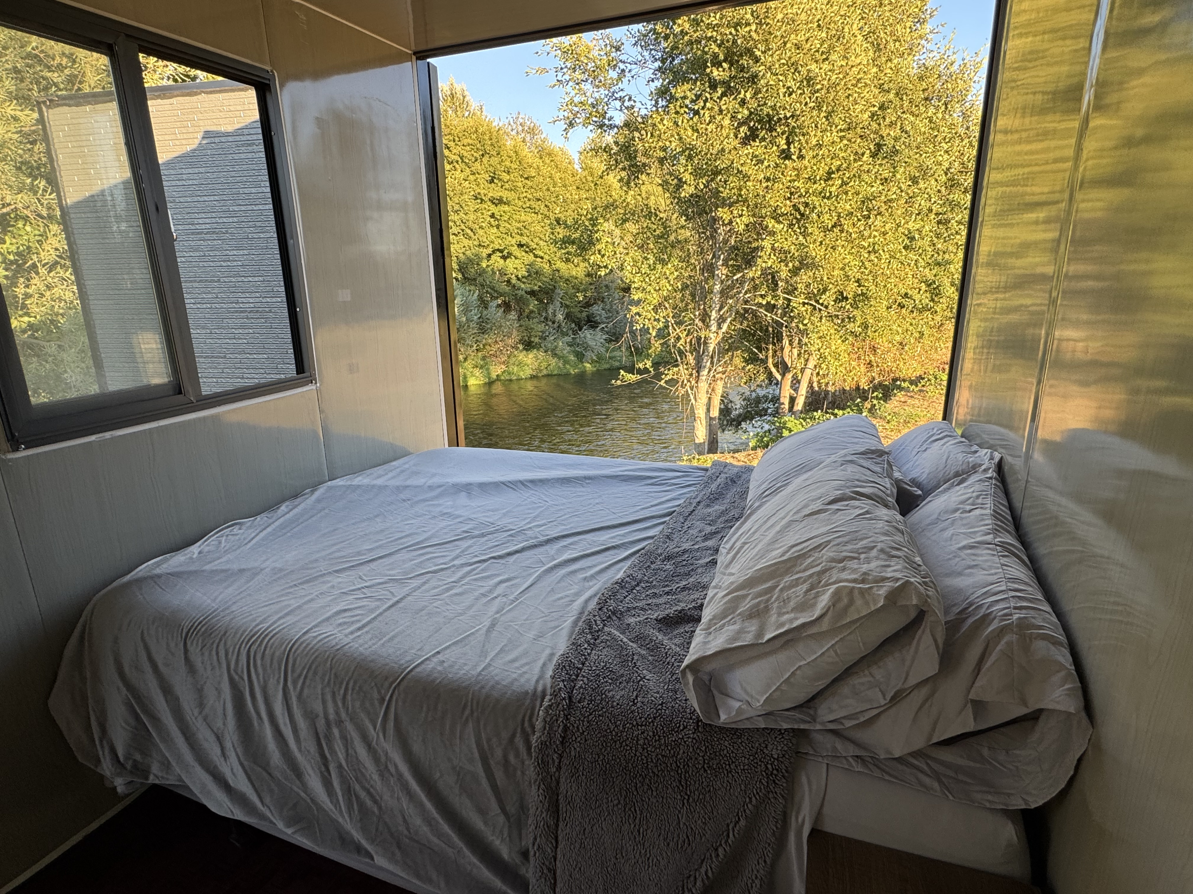RV rental bedroom with a cozy bed and panoramic river view. Large windows showcase lush trees and peaceful water.. Ford F350 2025