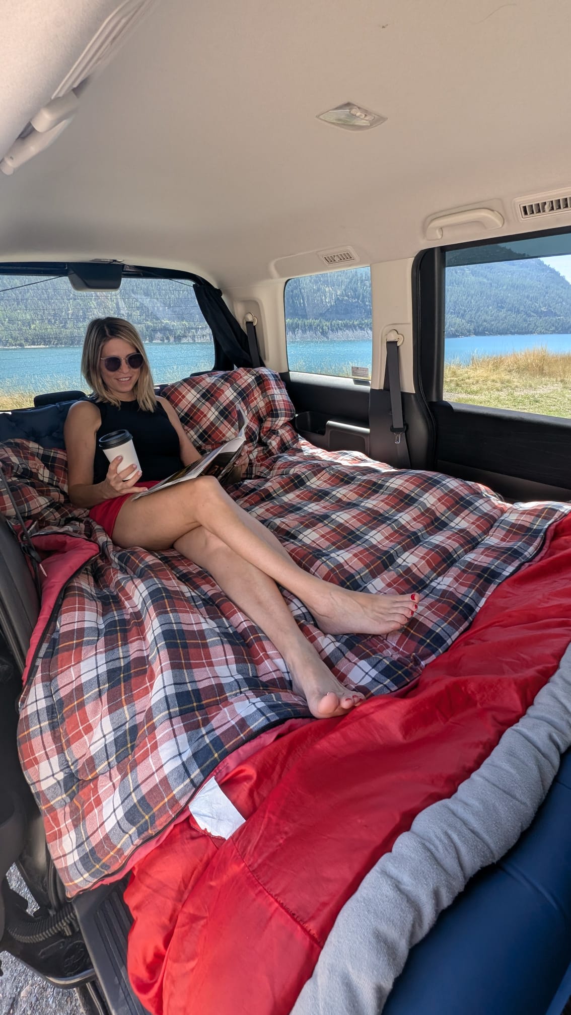 Relaxing inside a comfy **RV rental** with a great lake view. A smiling woman sips coffee & reads on a plaid bed.. Universal Transformer 2009