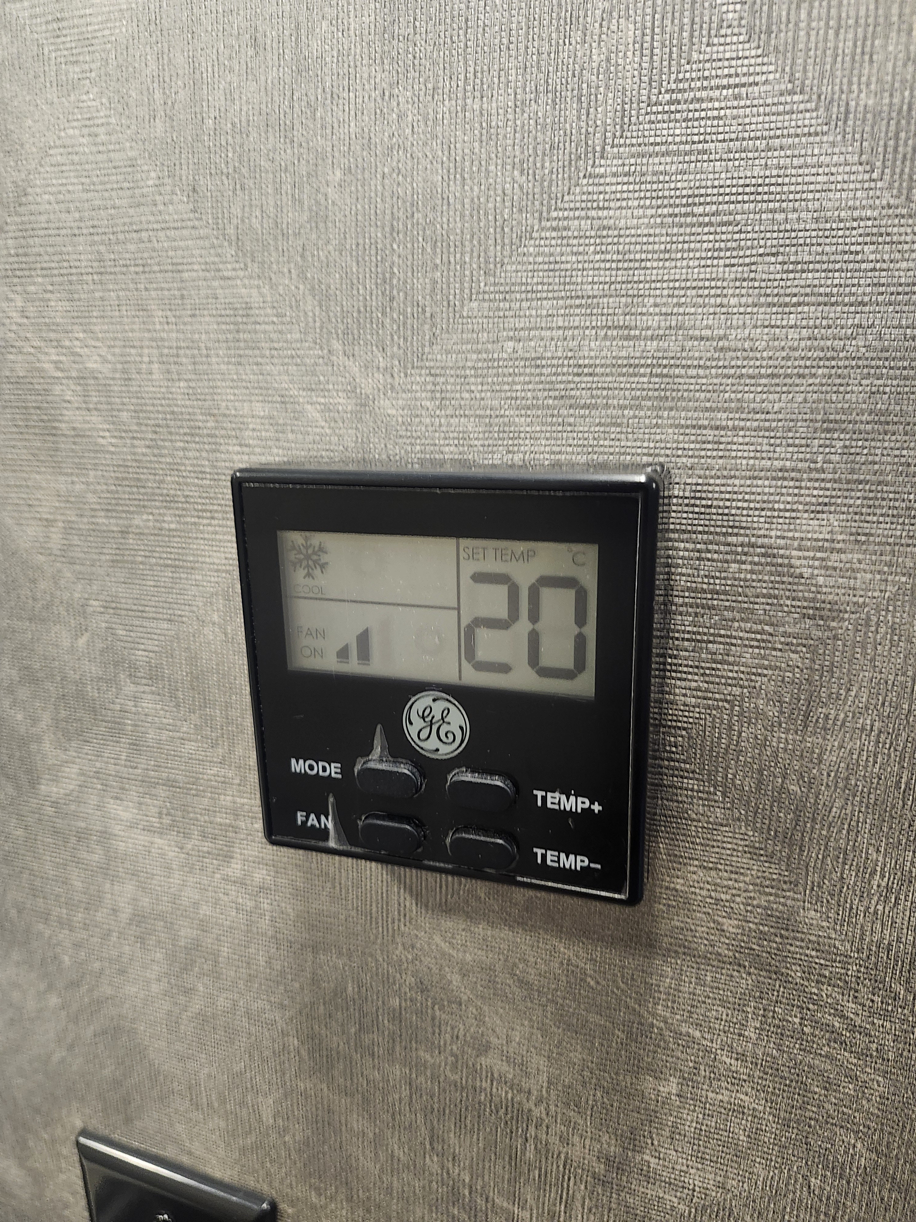 Black GE digital thermostat in an RV rental, set to 20°C (cool mode), fan on. Against a textured grey wall.. Forester LE 2023