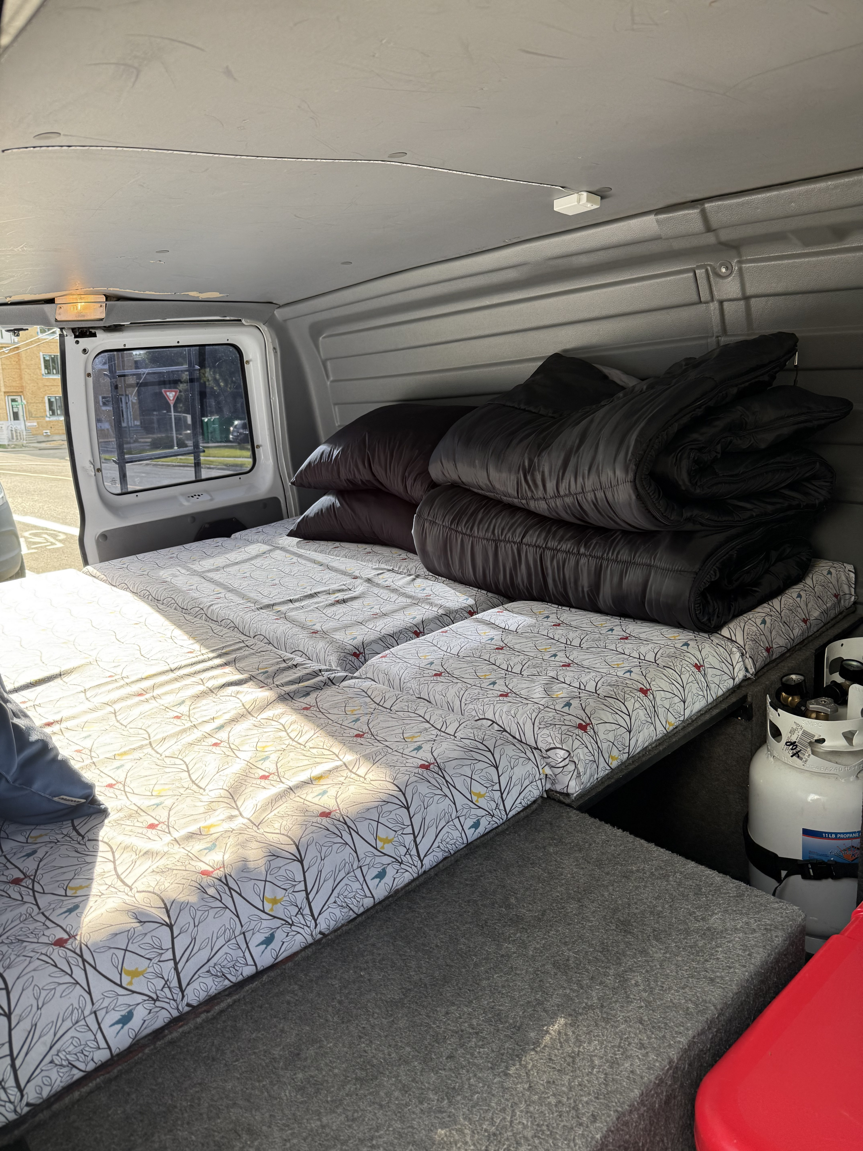 RV rental van interior: comfy bed with patterned mattresses, black sleeping bags, and a propane tank.. Ford Econoline 150 2005
