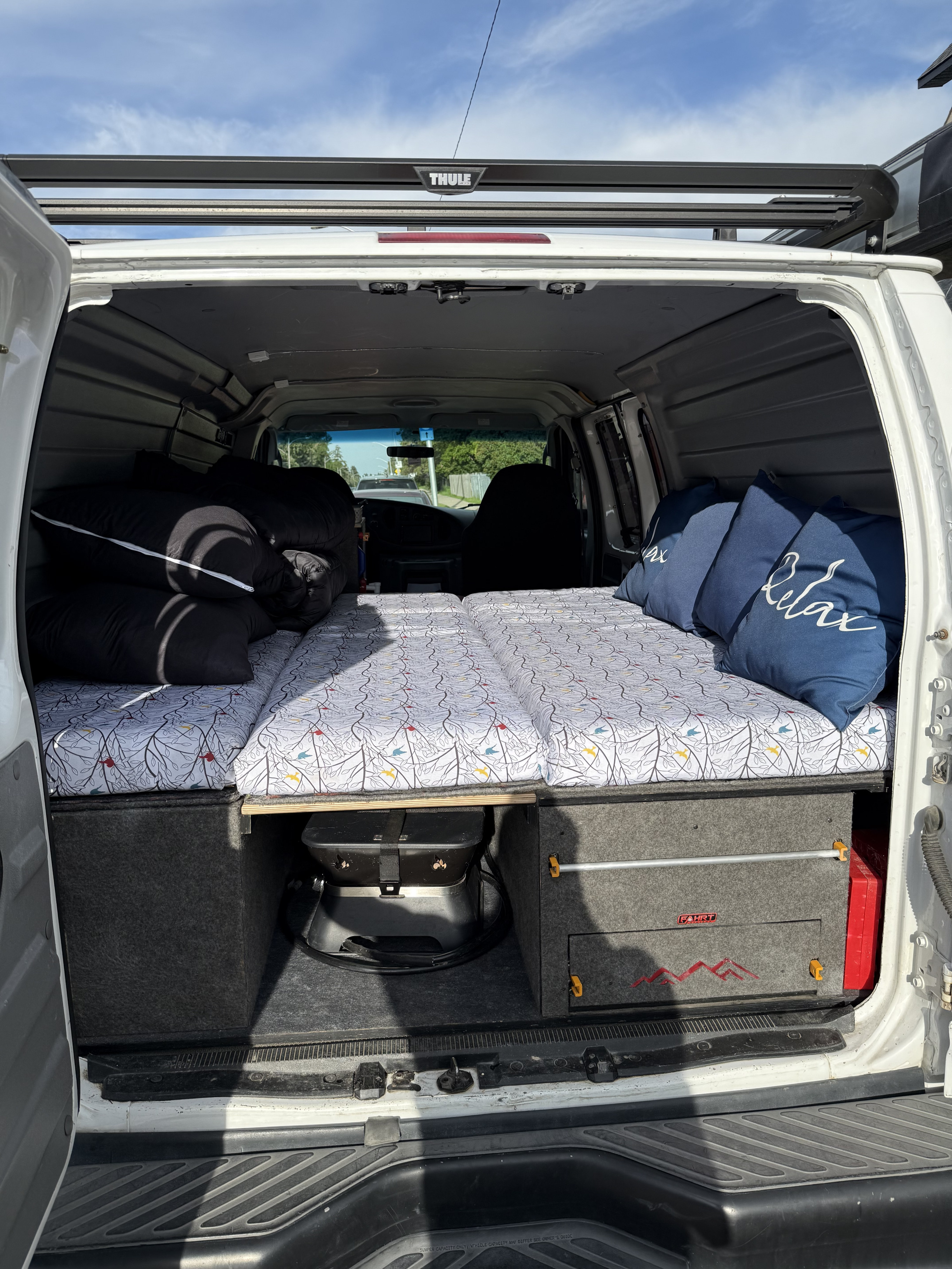 This RV rental offers a cozy, custom-built bed with "Relax" pillows, under-bed storage, and a visible Thule roof rack for travel.. Ford Econoline 150 2005