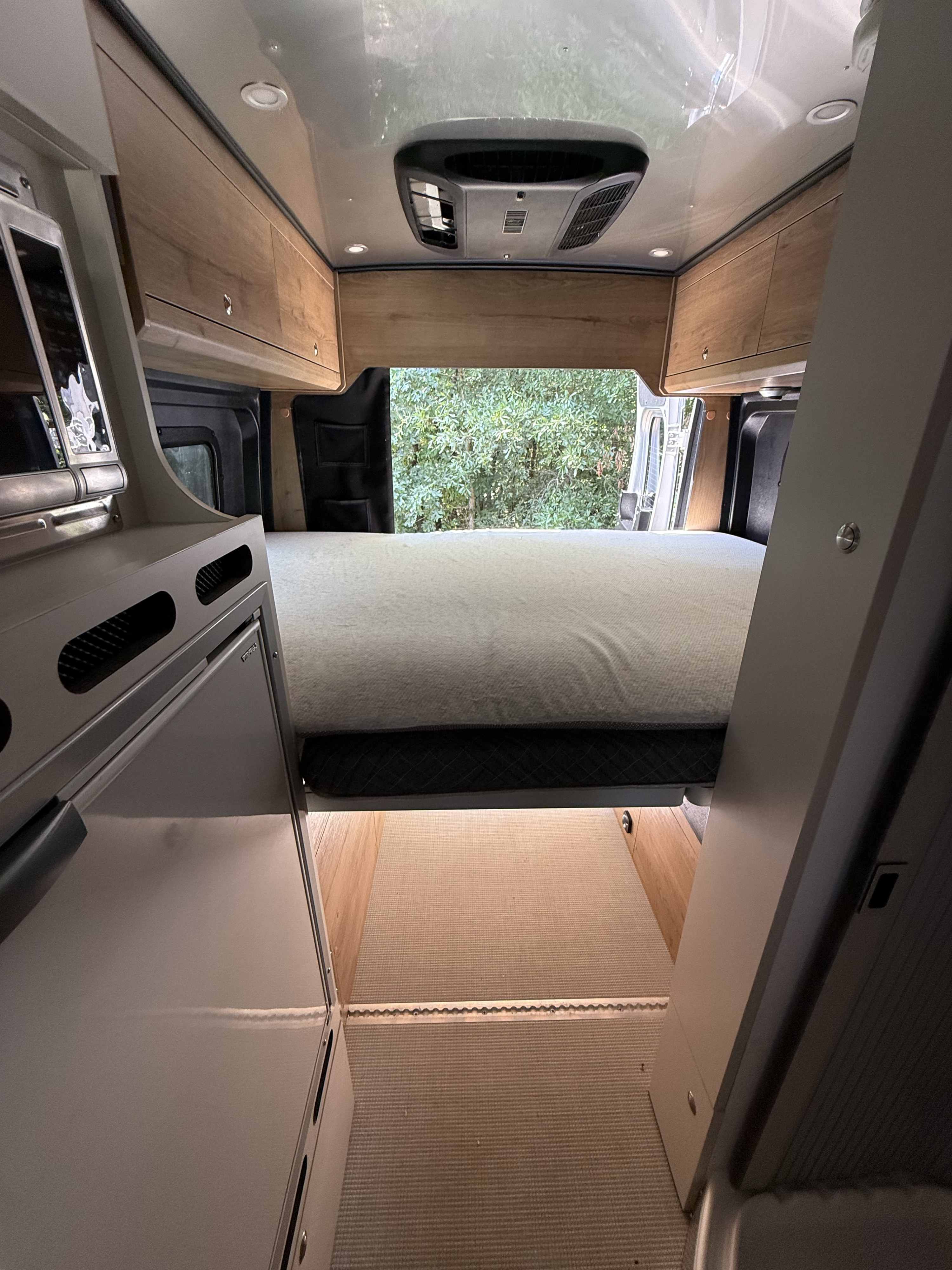 Inside a modern RV rental: view of the bed, overhead cabinets, AC, and rear opening to trees. Features a compact kitchen.. Airstream Airstream Class B 2023