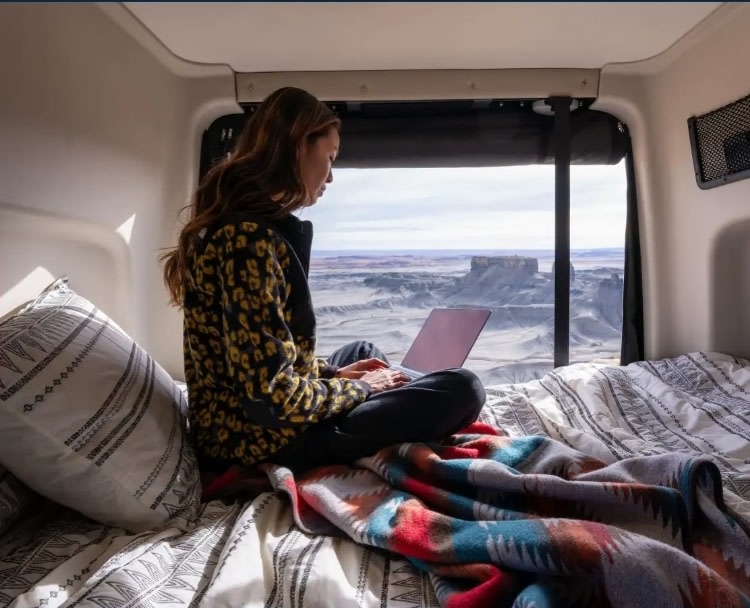 Enjoy remote work from an **RV rental**! A digital nomad uses a laptop inside a camper van with a stunning desert view.. Winnebago Revel 2025