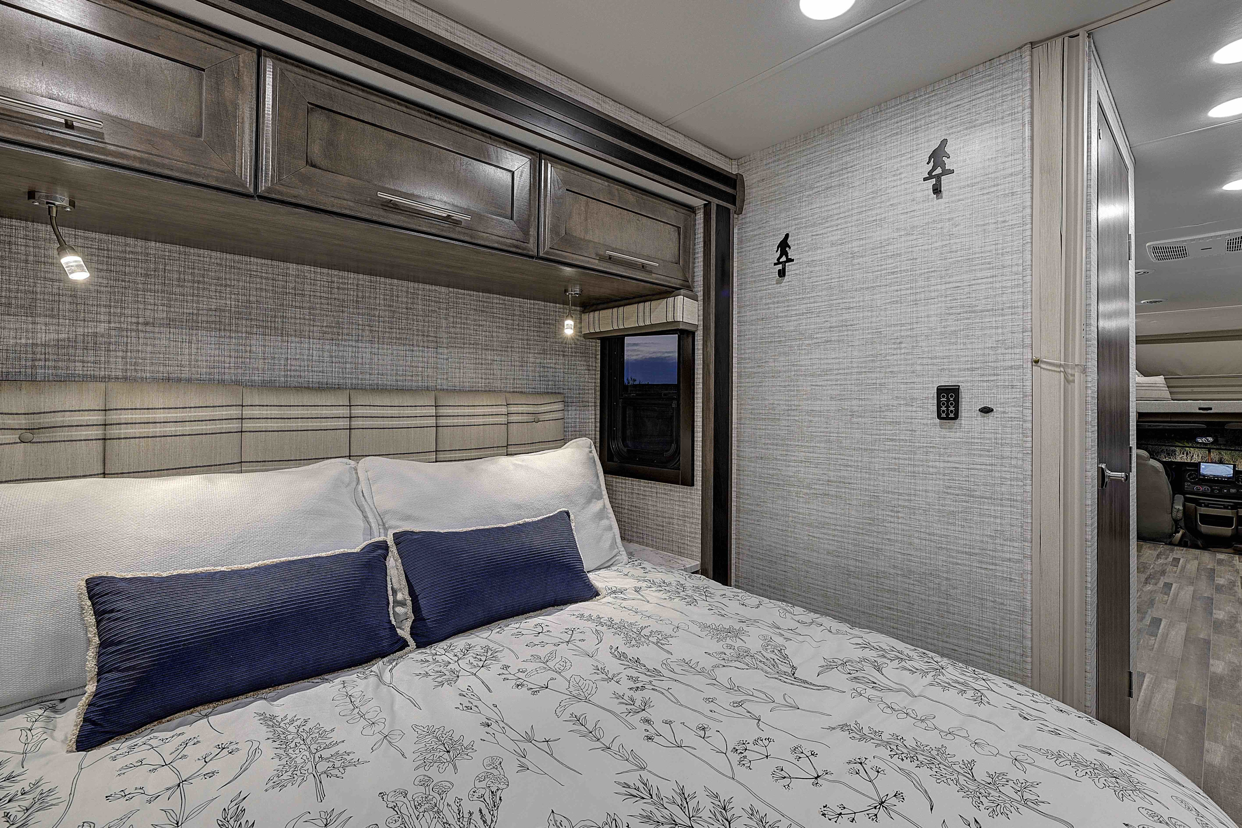 Relax in this RV rental bedroom with a comfortable bed, overhead cabinets, reading lights, and fun Bigfoot wall hooks.. Entegra Esteem 2025