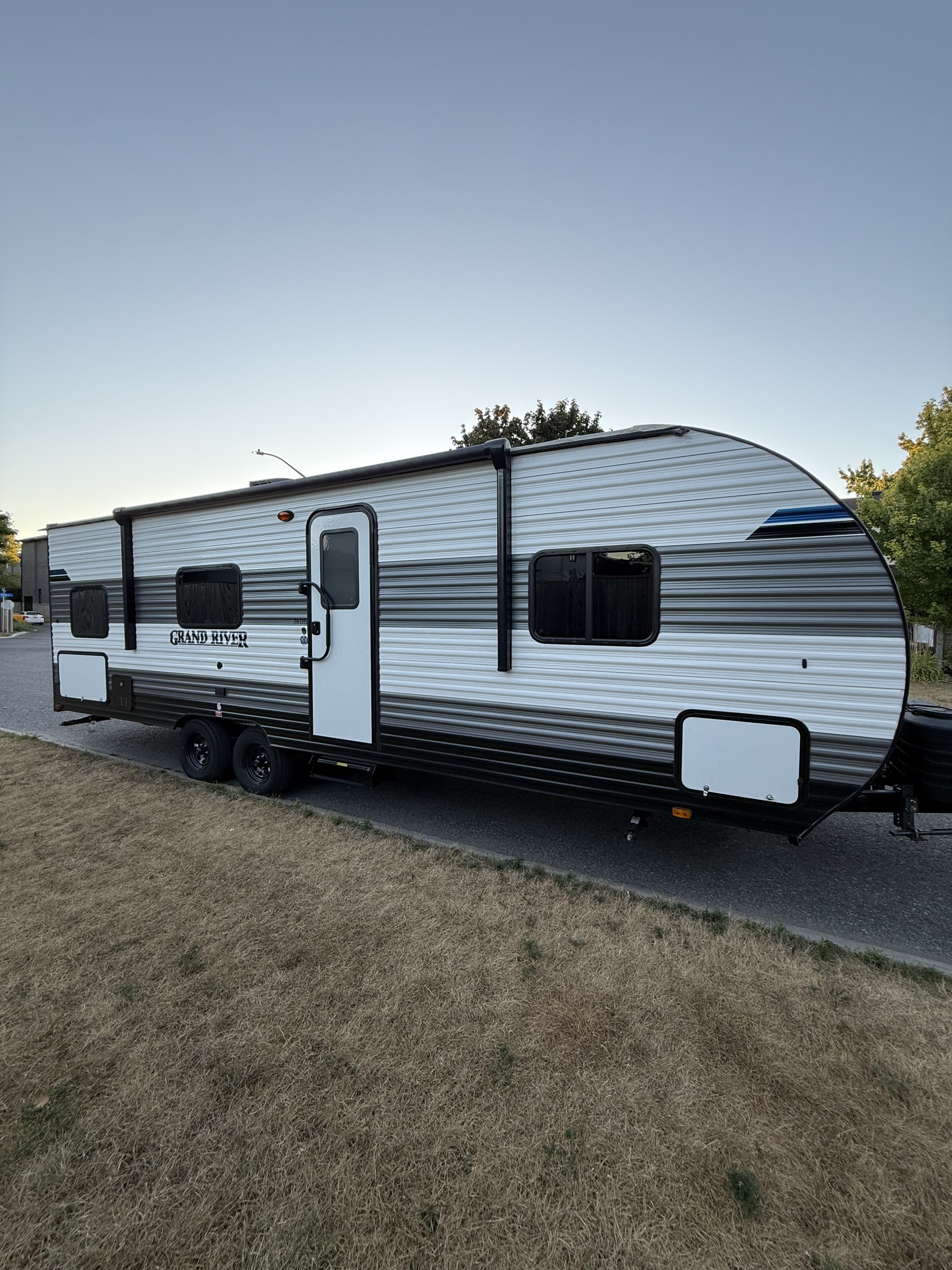 This white and gray "Grand River" travel trailer, perfect for rv rental, is parked on asphalt beside dry grass under a clear sky.. Gulf Stream Grand river 2026