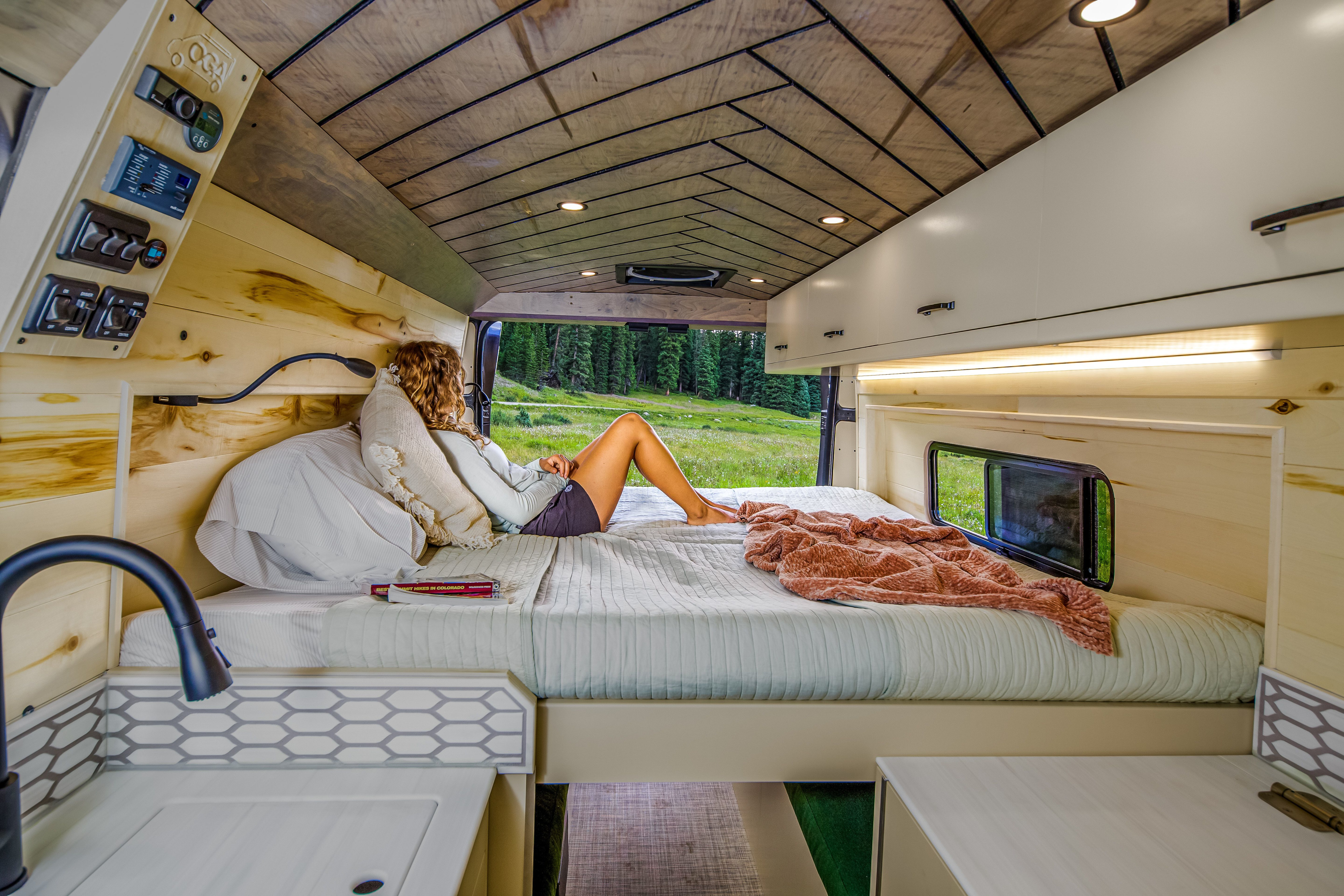 Relaxing in an rv rental camper van, a woman on a bed enjoys a forest view. Modern wood interior with controls & kitchen sink.. RAM Promaster 2500 2021