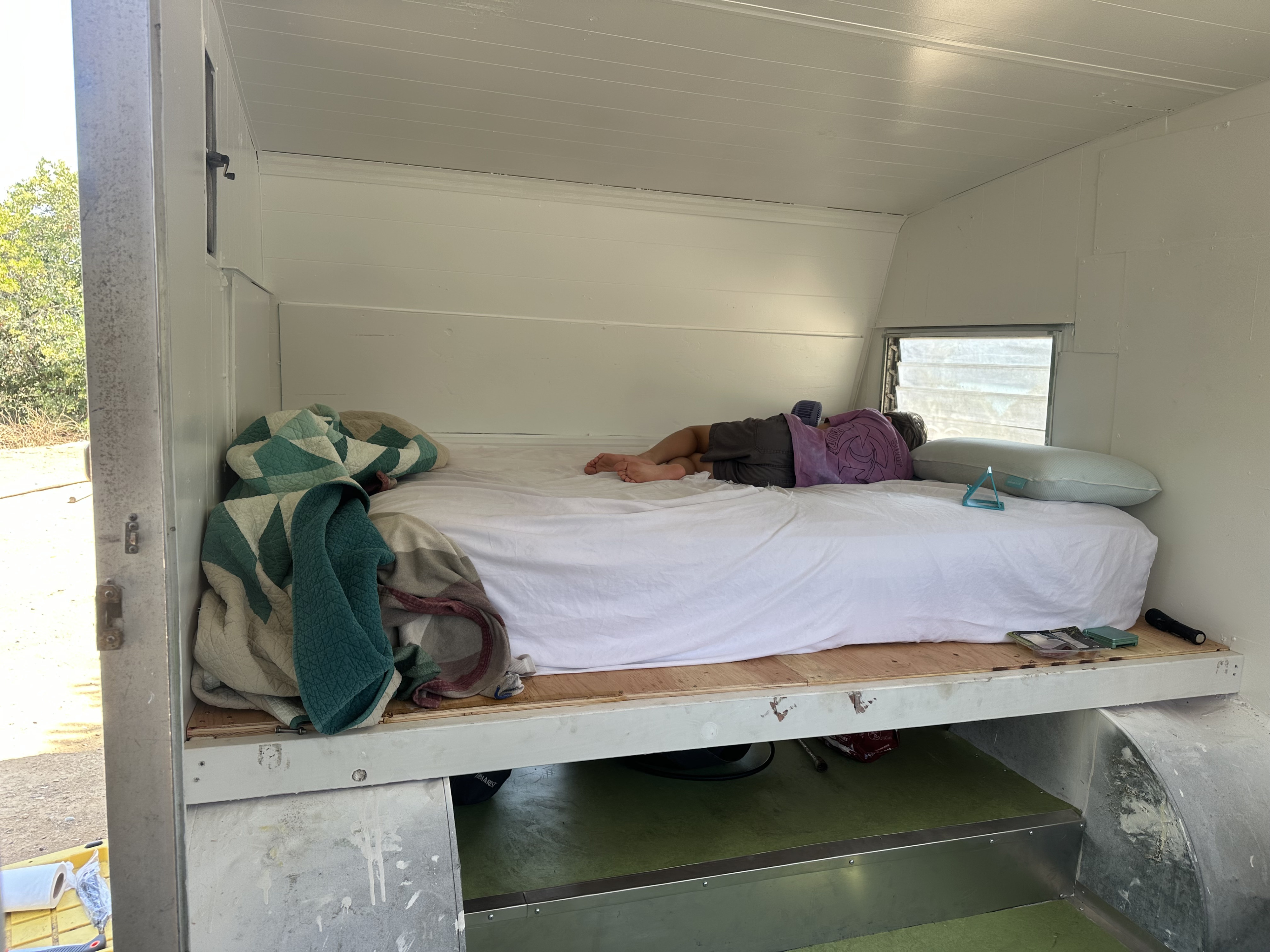 Cozy RV rental interior: A person naps on a bed with blankets and a pillow, showcasing the comfortable sleeping setup in a converted trailer.. Aristocrat Lo-Liner 1965