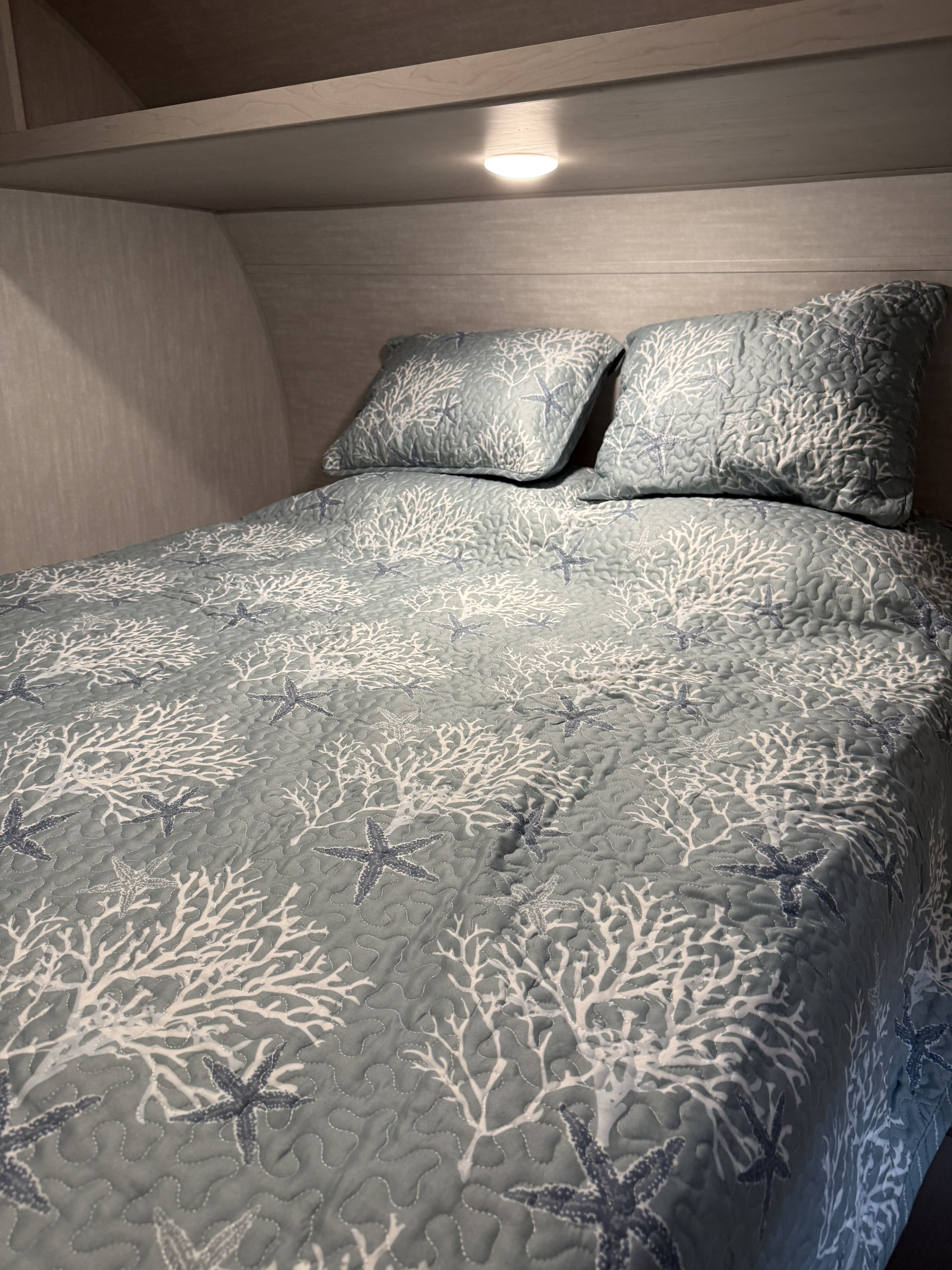 Inside an RV rental: a comfy bedroom with a blue coastal quilt, featuring white coral & starfish, and an overhead light.. Coachmen Clipper 25BH 2025