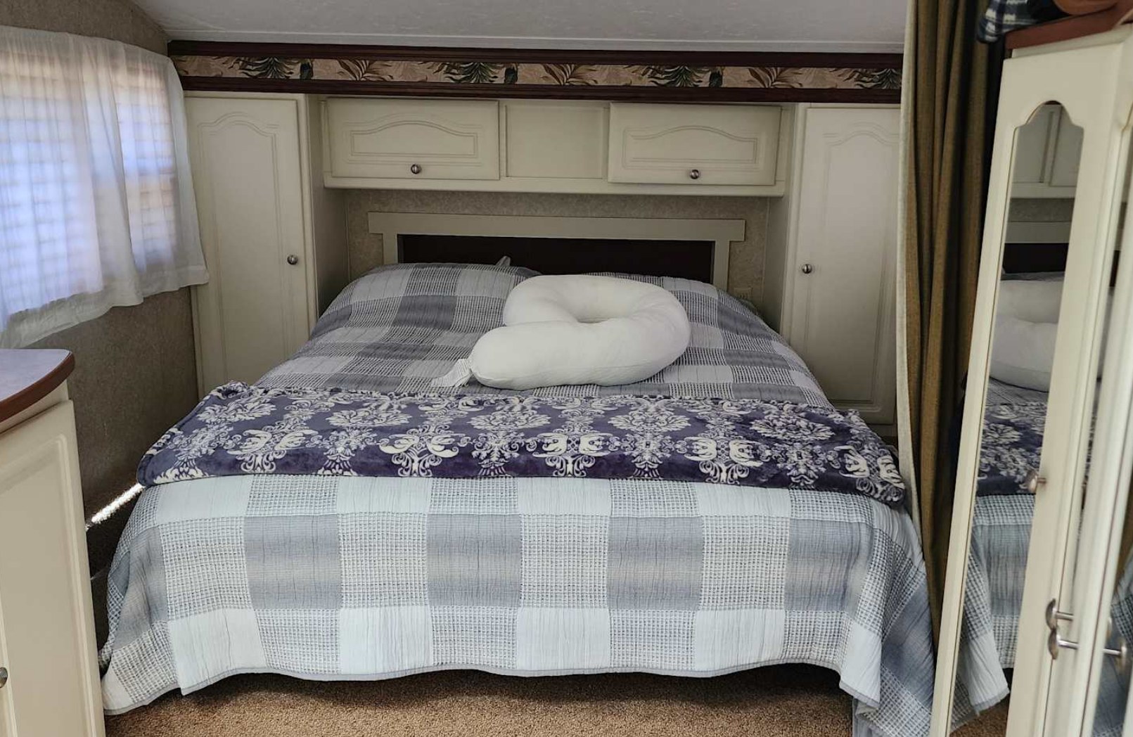 A cozy RV rental bedroom features a comfortable bed with checkered and floral blankets, a large U-shaped pillow, overhead cabinets, and a window for natural light.. Keystone RV Outback Sydney 2007
