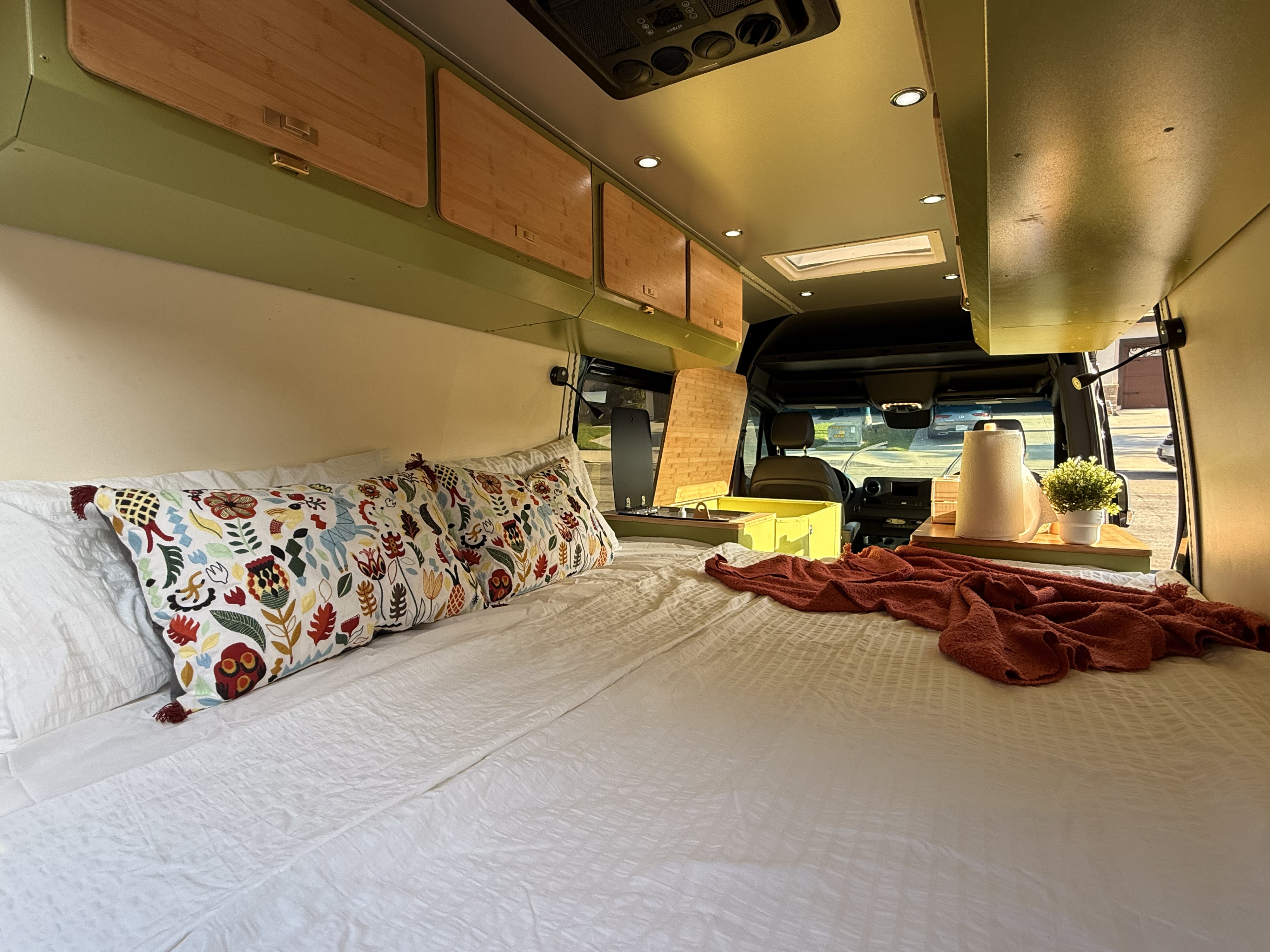 Cozy RV rental interior with a comfortable bed, vibrant pillows, olive green overhead cabinets, and a compact kitchen area.. MERCEDES BENZ SPRINTER 2020