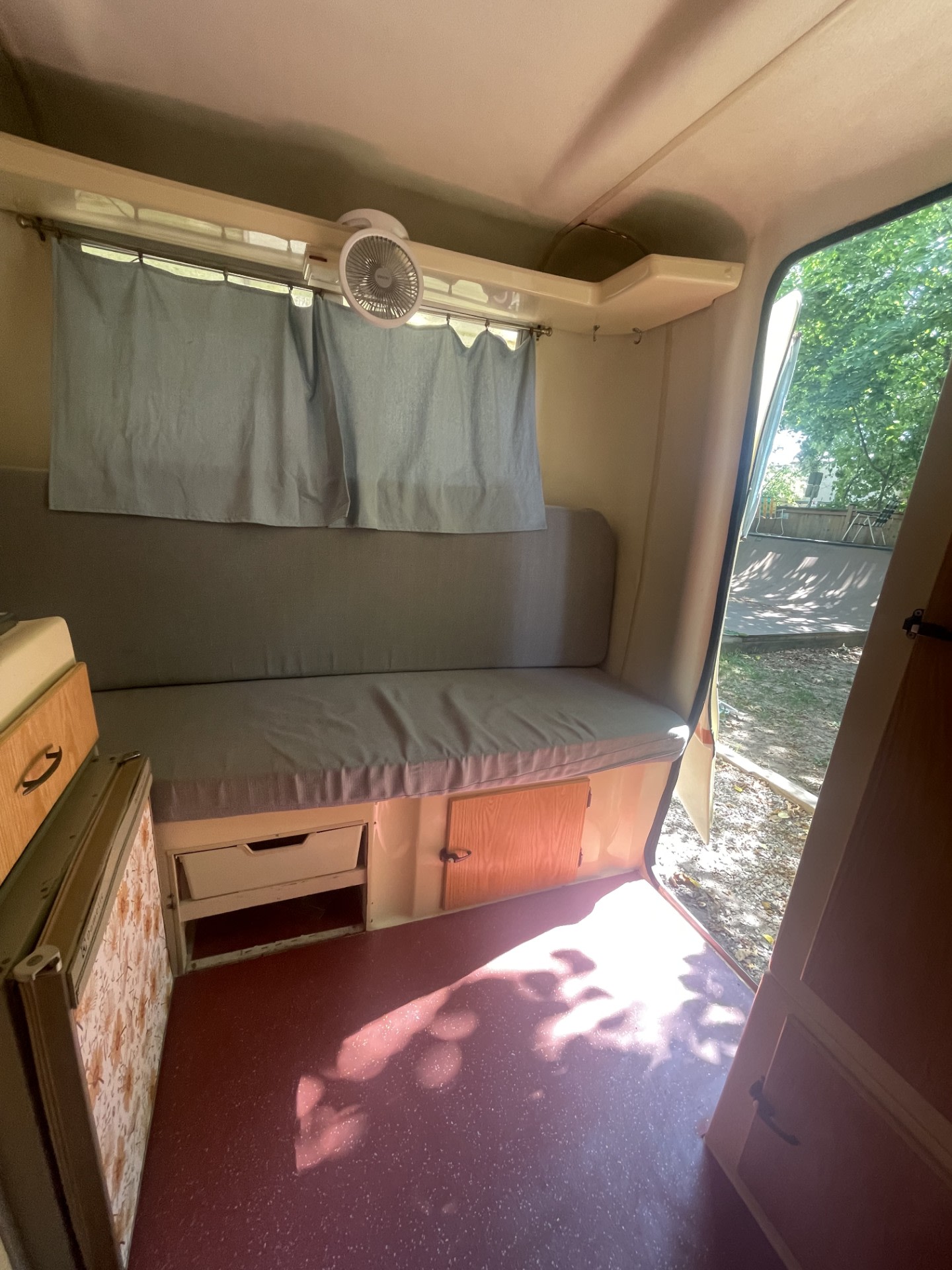 Compact RV rental interior with bench seat, fan, and storage, looking out to nature.. Trillium Rv 130 1976