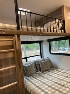 This RV rental interior features a comfortable lower bed with plaid bedding, an accessible upper bunk with a safety railing, and a ladder.. Keystone RV Arcadia 2022