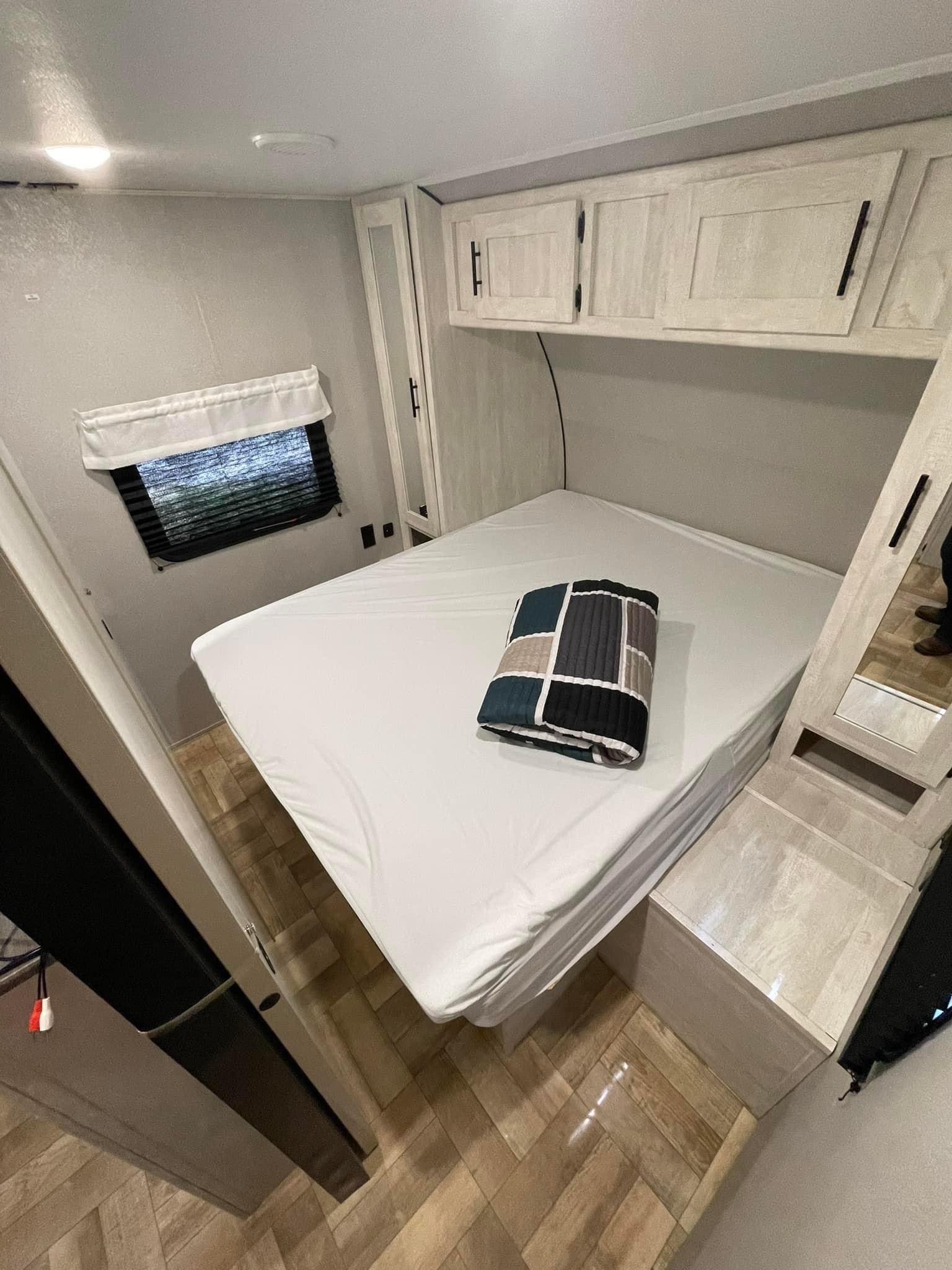 Inside an RV rental, a compact bedroom features a white bed, patterned blanket, overhead cabinets, window, and mirrored wardrobe.. Forest River Shasta 2022