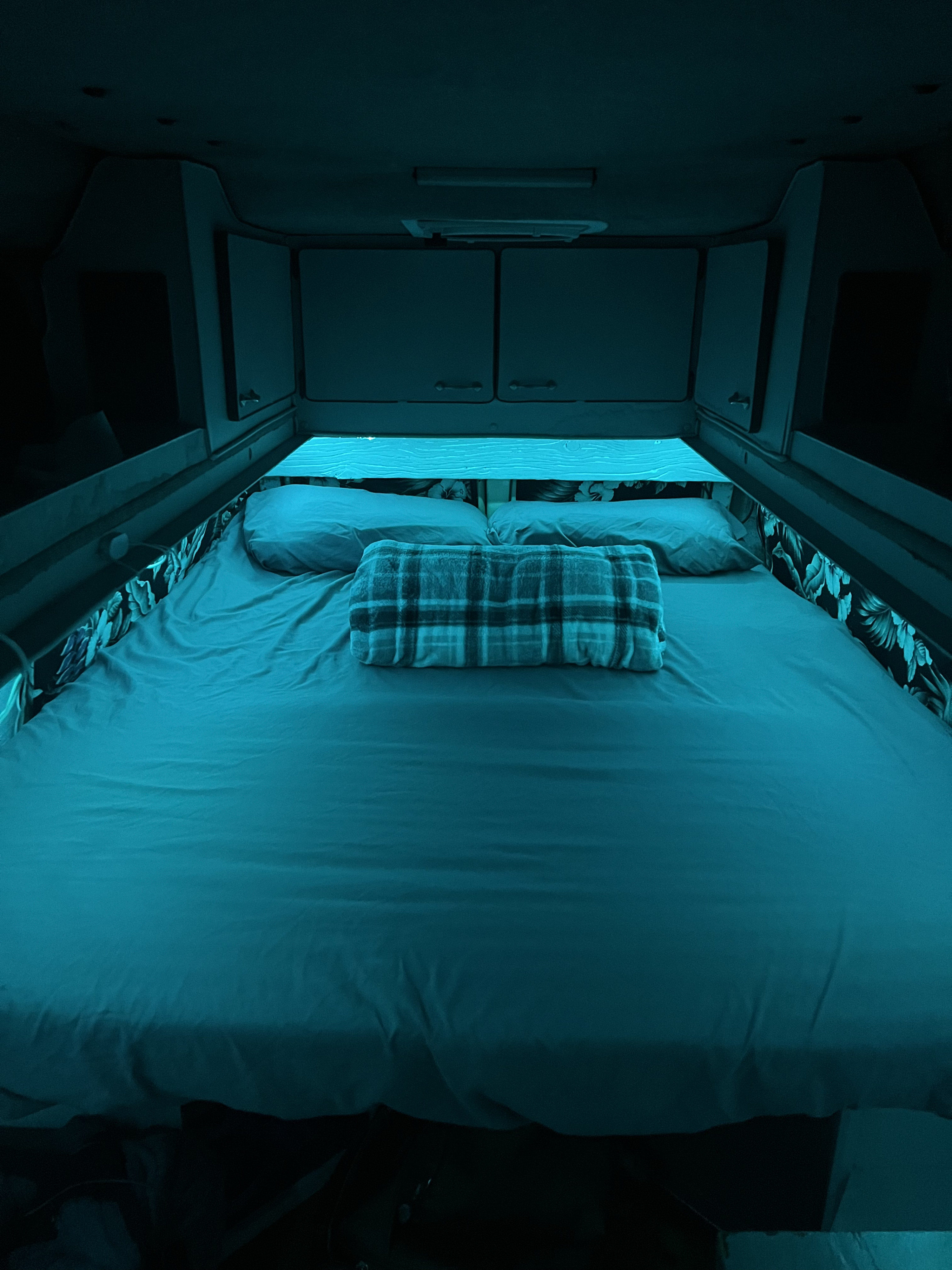RV rental's cozy bedroom with a large bed, blue ambient lighting, and overhead cabinets.. Dodge B Van 1994
