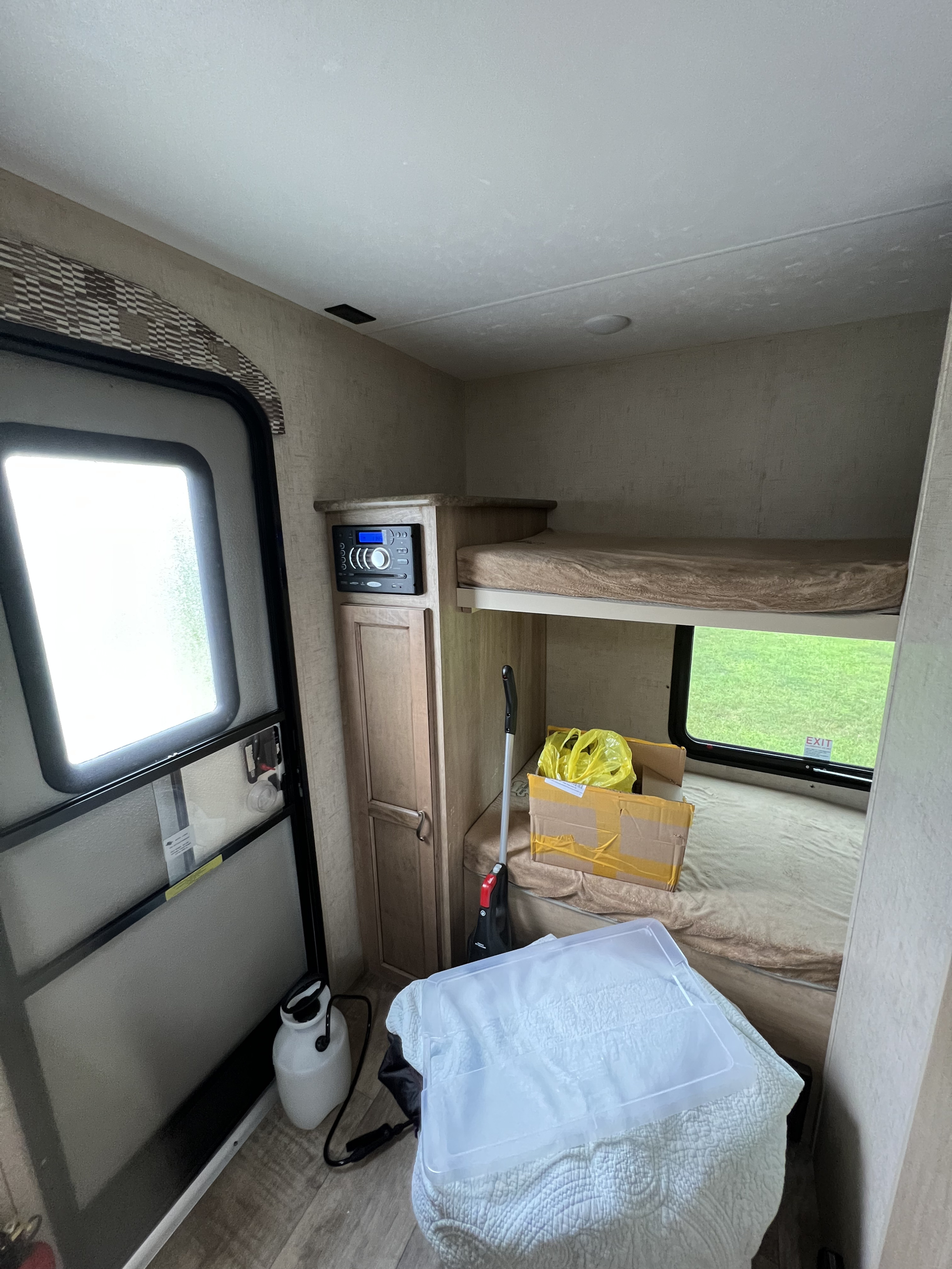Interior of an RV rental: bunk beds, door, and storage with cleaning tools & boxes. Green outside window.. KZ VISION 2017