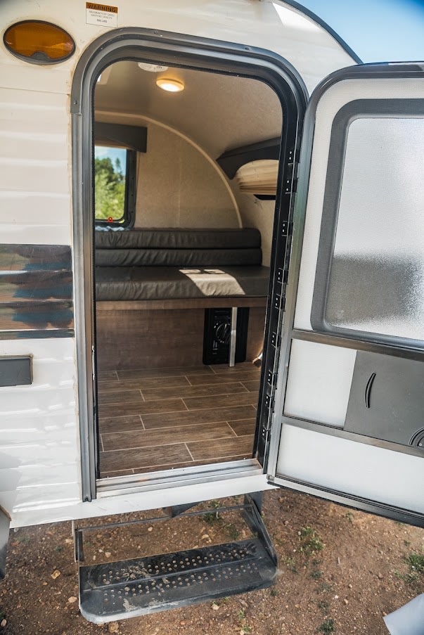 Open door of an RV rental revealing its compact interior with seating, window, and wood-look flooring, showing the entrance steps.. Sunset Park RV SunRay 2021