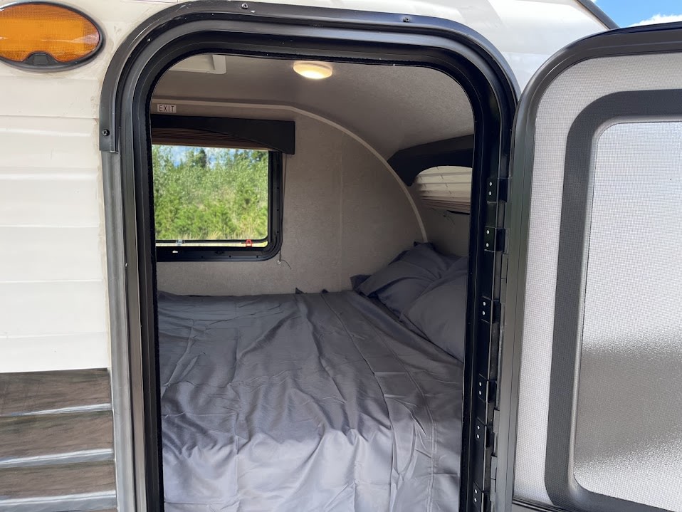 Cozy rv rental interior: a comfy bed with grey sheets, ceiling light, and window revealing green trees outside.. Sunset Park RV SunRay 2021