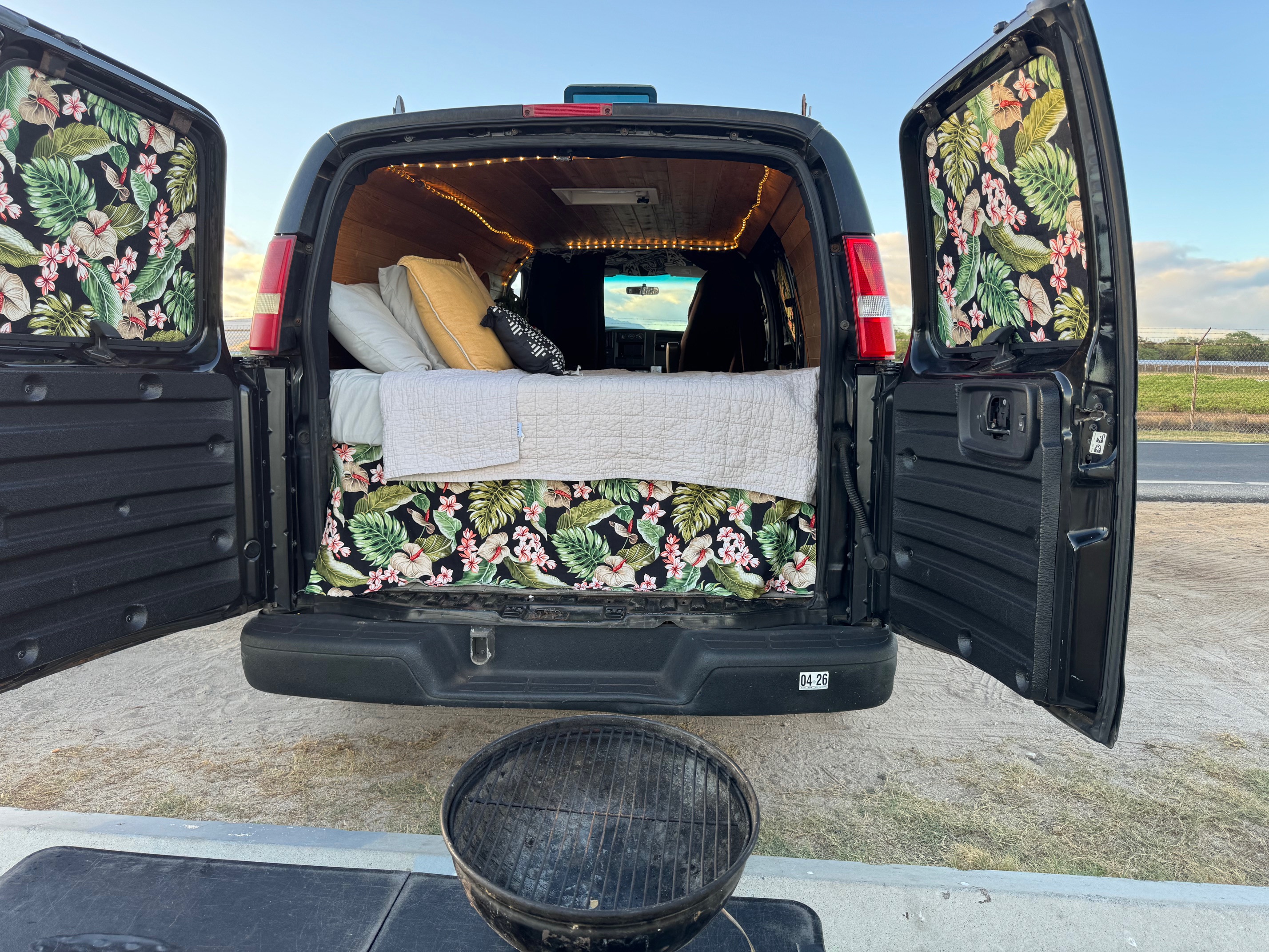 Inside an open **rv rental** camper van: bed with tropical decor, string lights, and grill setup for outdoor adventure.. Chevrolet Express 2004