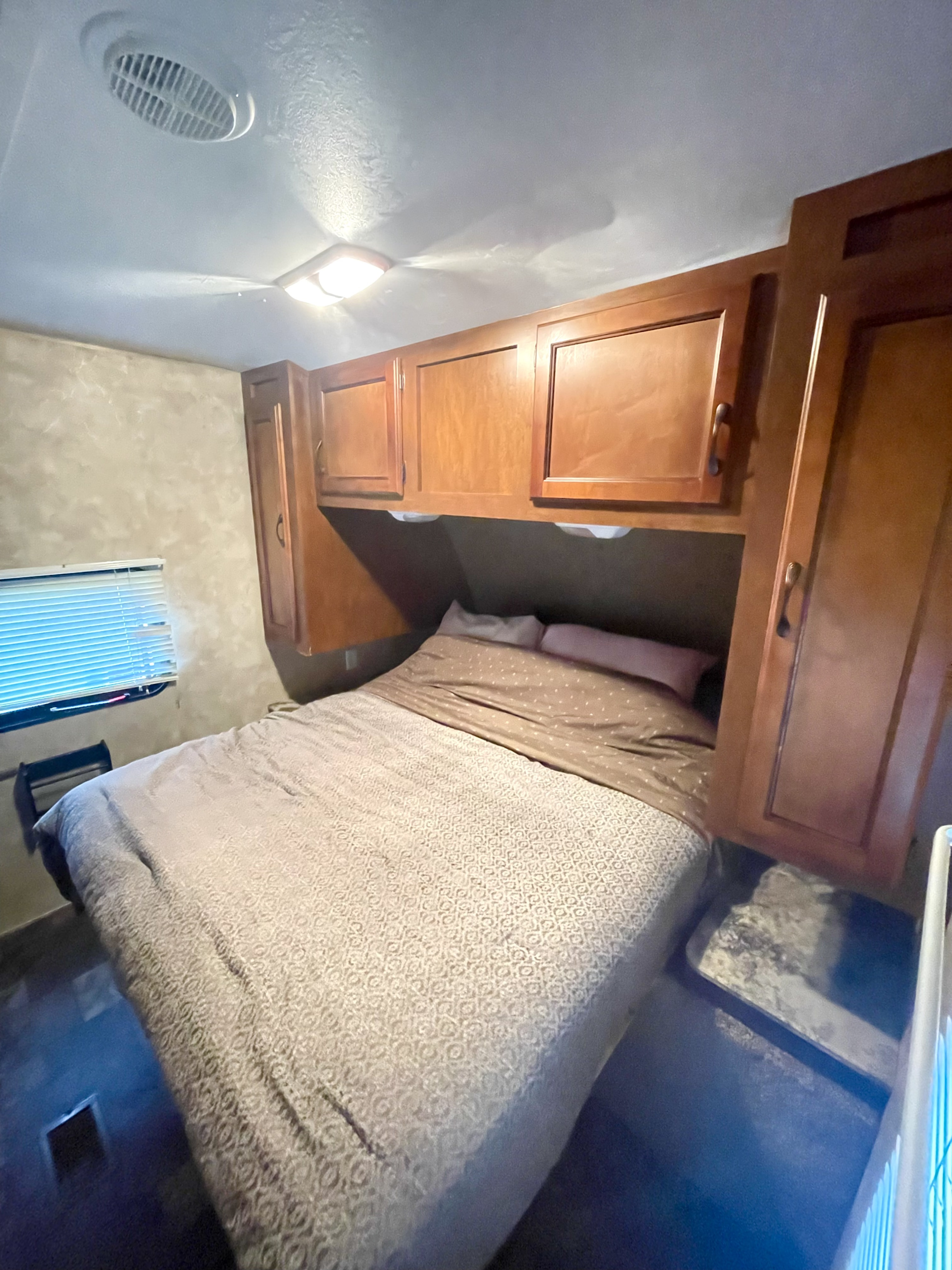 Experience an RV rental: a compact bedroom with a patterned bed, overhead cabinets, a window, and ceiling vent/light.. Coachmen Catalina Deluxe Edition 2013