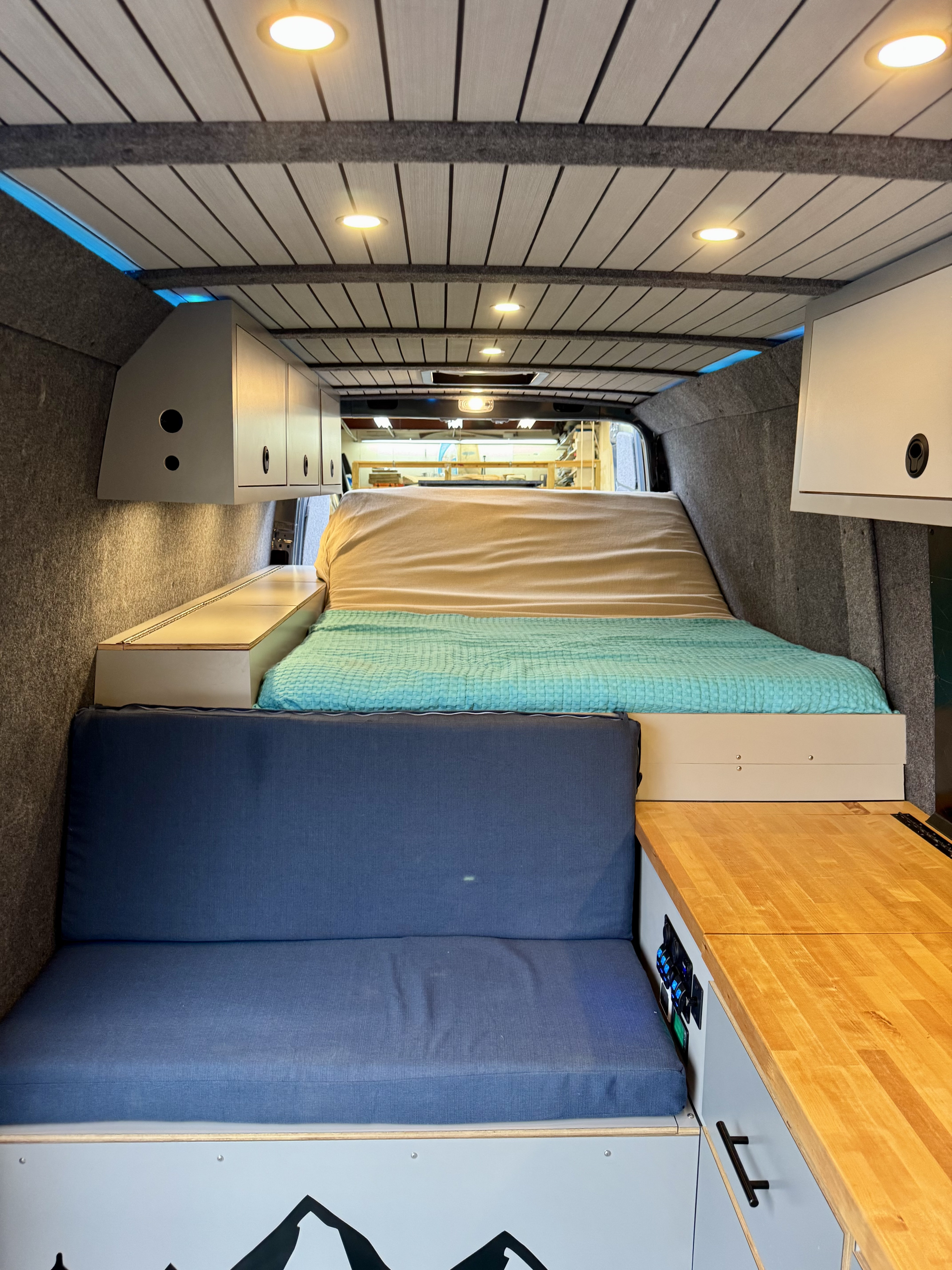 This well-appointed **RV rental** van interior showcases a bed, a blue cushioned bench, wooden countertop, and ample storage, lit by warm ceiling lights.. Mercedes Sprinter 2021