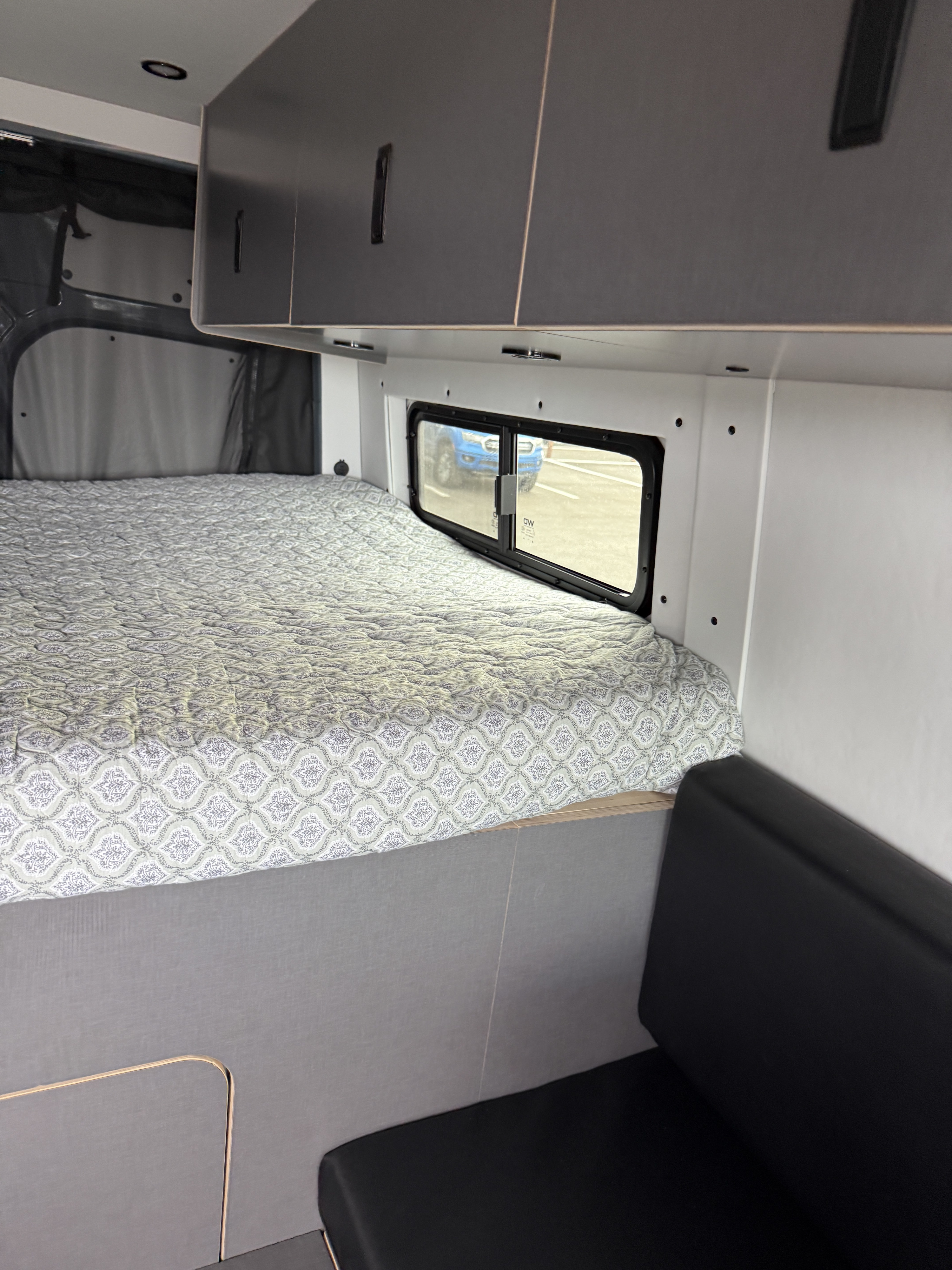 RV rental interior: cozy bed with patterned quilt, overhead cabinets, window with outside view, and black bench seat.. Mercedes Sprinter 2024