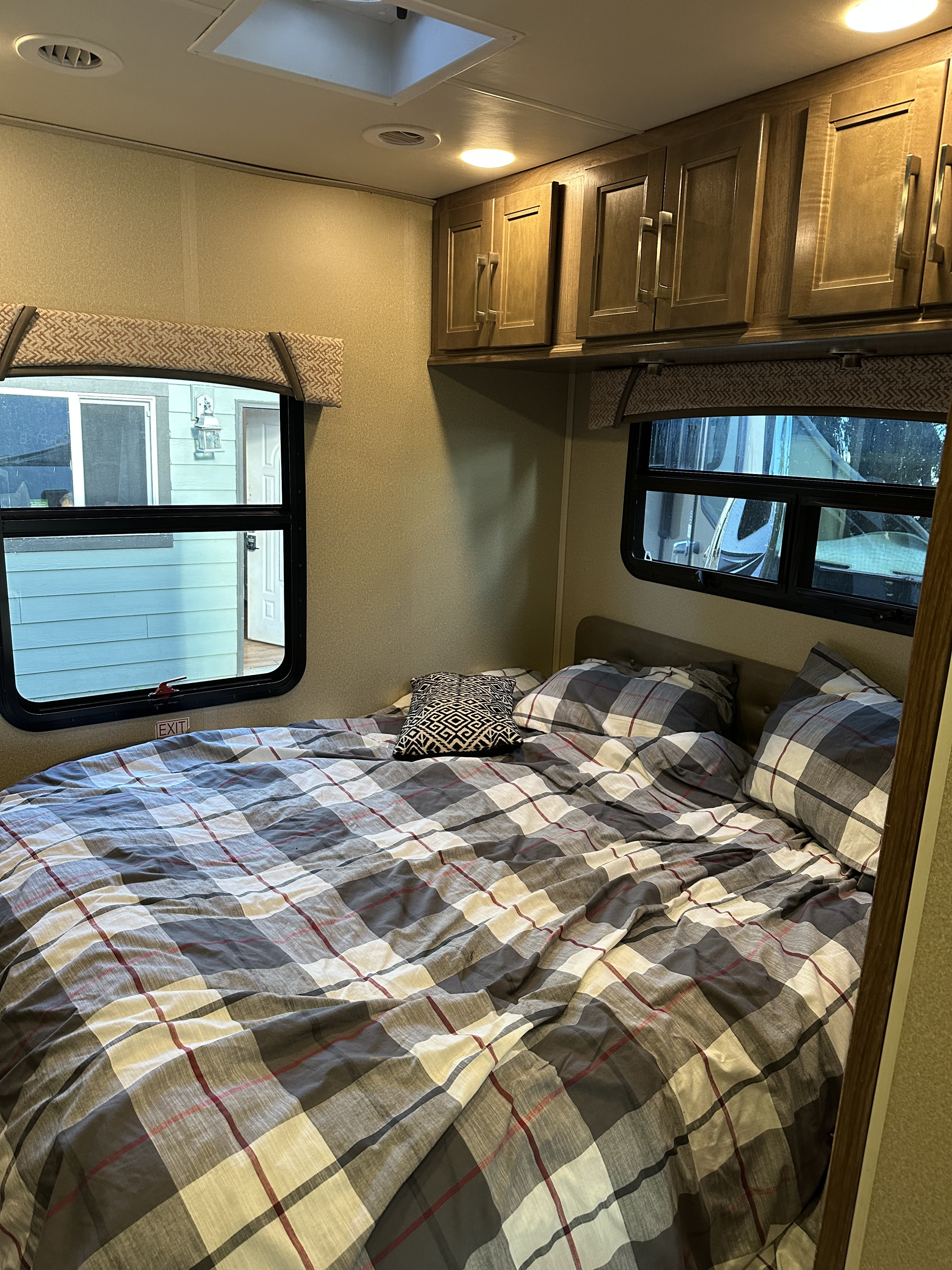 An RV rental bedroom with a plaid bed, an "EXIT" sign, overhead cabinets, and windows revealing outdoor views.. Jayco Greyhawk 2017