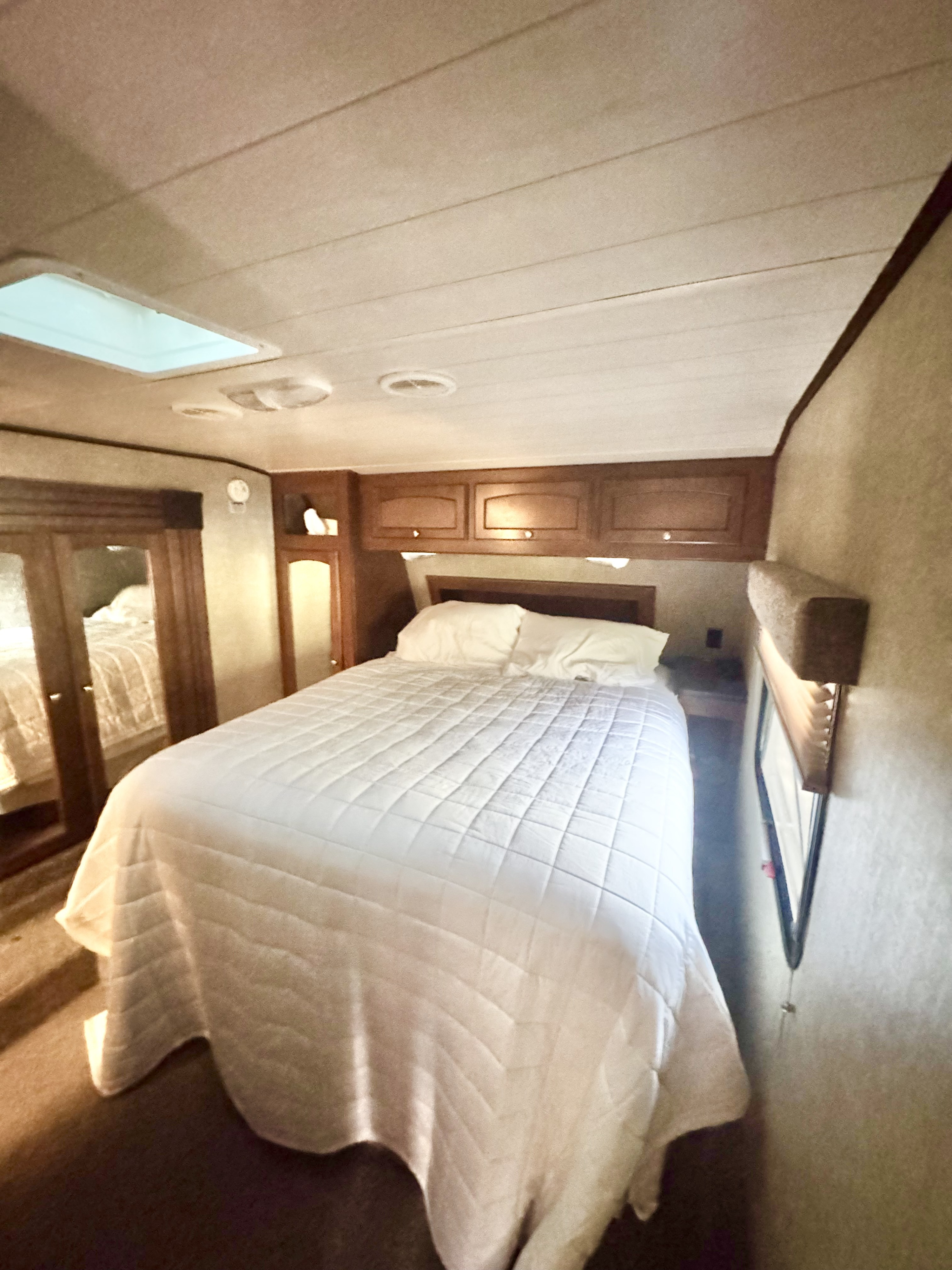 Well-appointed RV rental bedroom with a bed, white bedding, wooden overhead cabinets, a mirrored closet, and ceiling skylight.. Heartland RVs Global nomad 2020