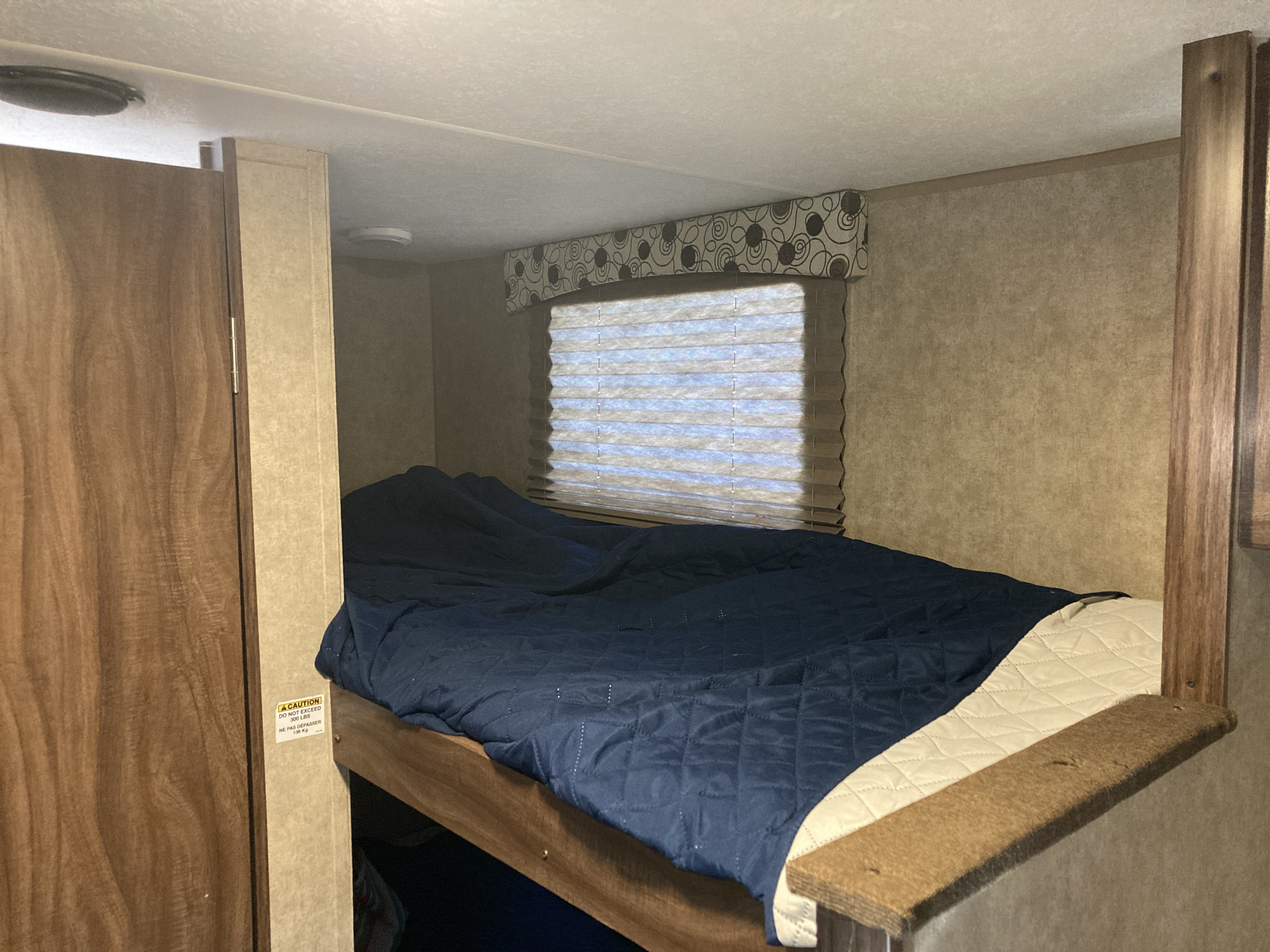 RV rental bunk bed: a cozy sleeping area with a dark blue comforter, light beige quilted blanket, and a window with blinds.. Clipper 17BH 2018