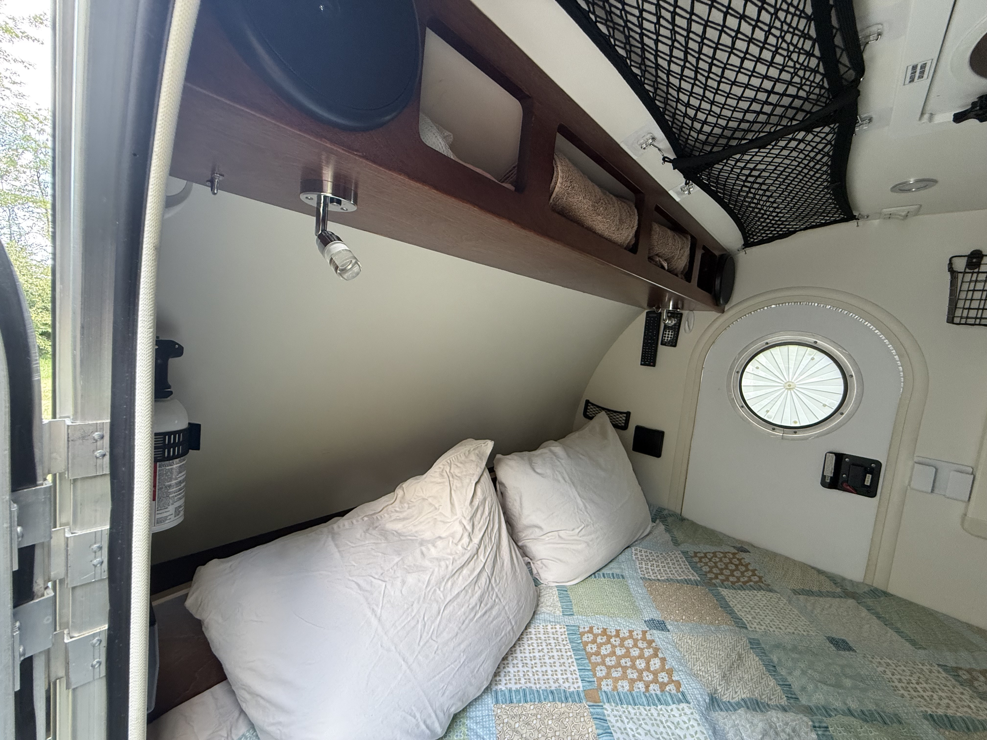 Cozy RV rental bedroom showing a bed with a quilt, overhead storage, reading lights, a round window, and a visible fire extinguisher.. Little Guy T@G 2016