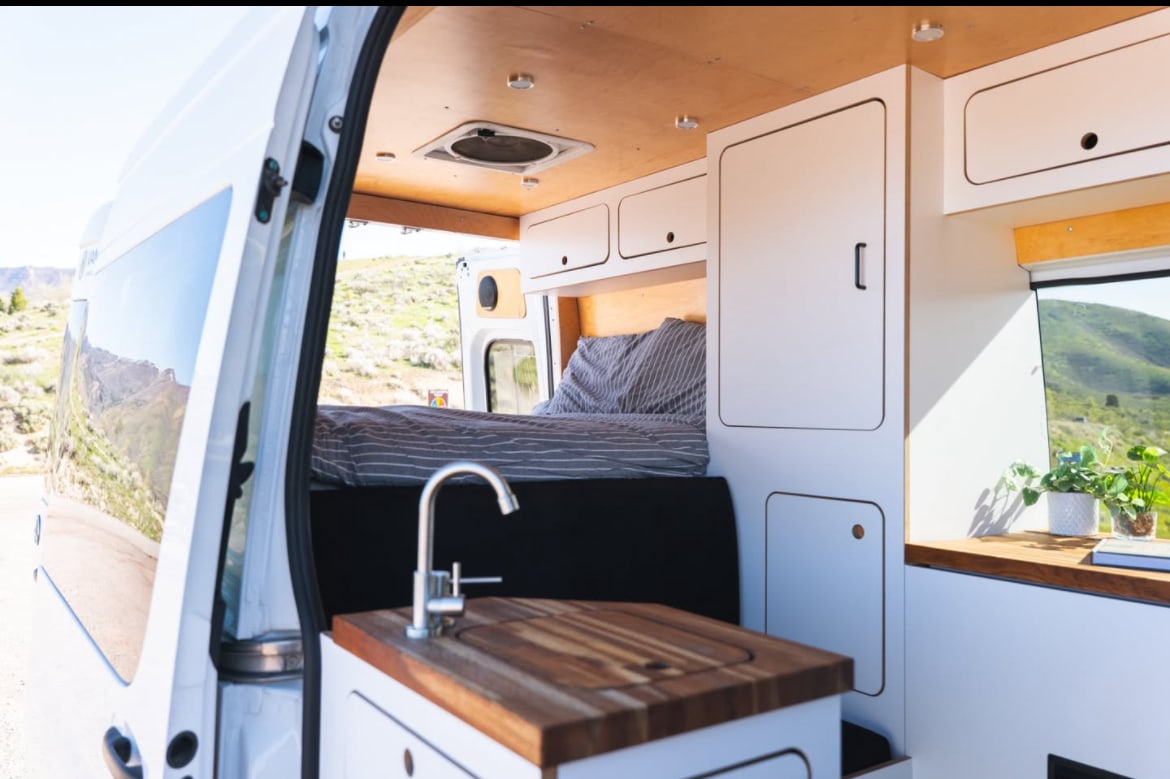 Step inside this RV rental: a cozy bed, compact kitchen with wooden counter/sink, & white storage cabinets for your van life journey.. Mercedes-Benz Sprinter RV Motorhome Campervan 2022