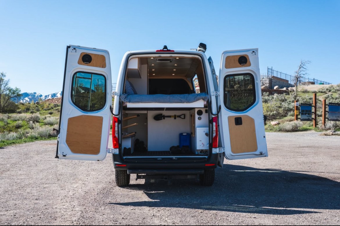 Rear view of an rv rental: a white camper van with open doors, showing a bed platform & organized storage for adventure.. Mercedes-Benz Sprinter 2022