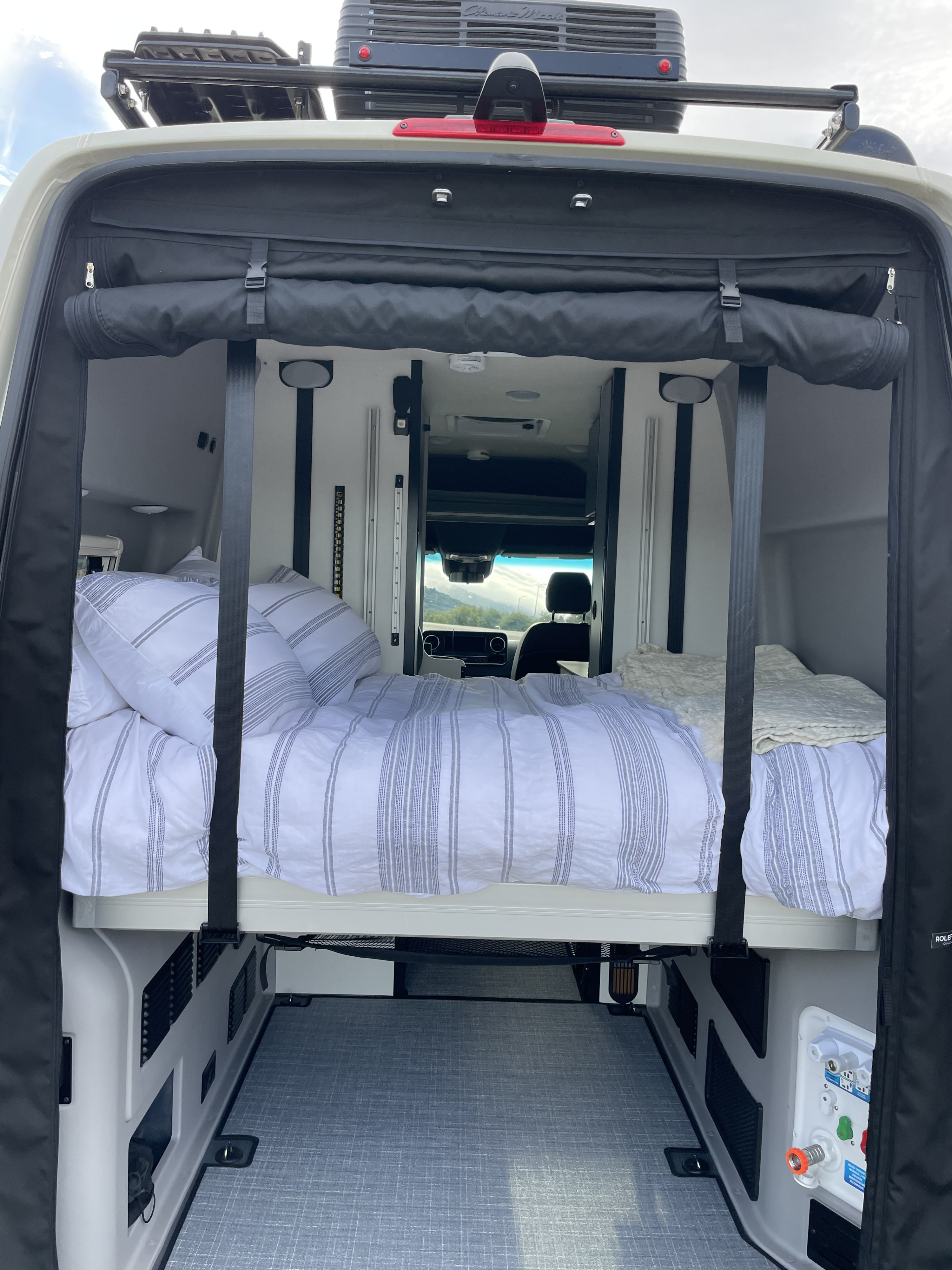 Inside an RV rental camper van: elevated bed with striped bedding, utility connections, and rear storage area, ideal for mobile living.. Winnebago Revel 2023