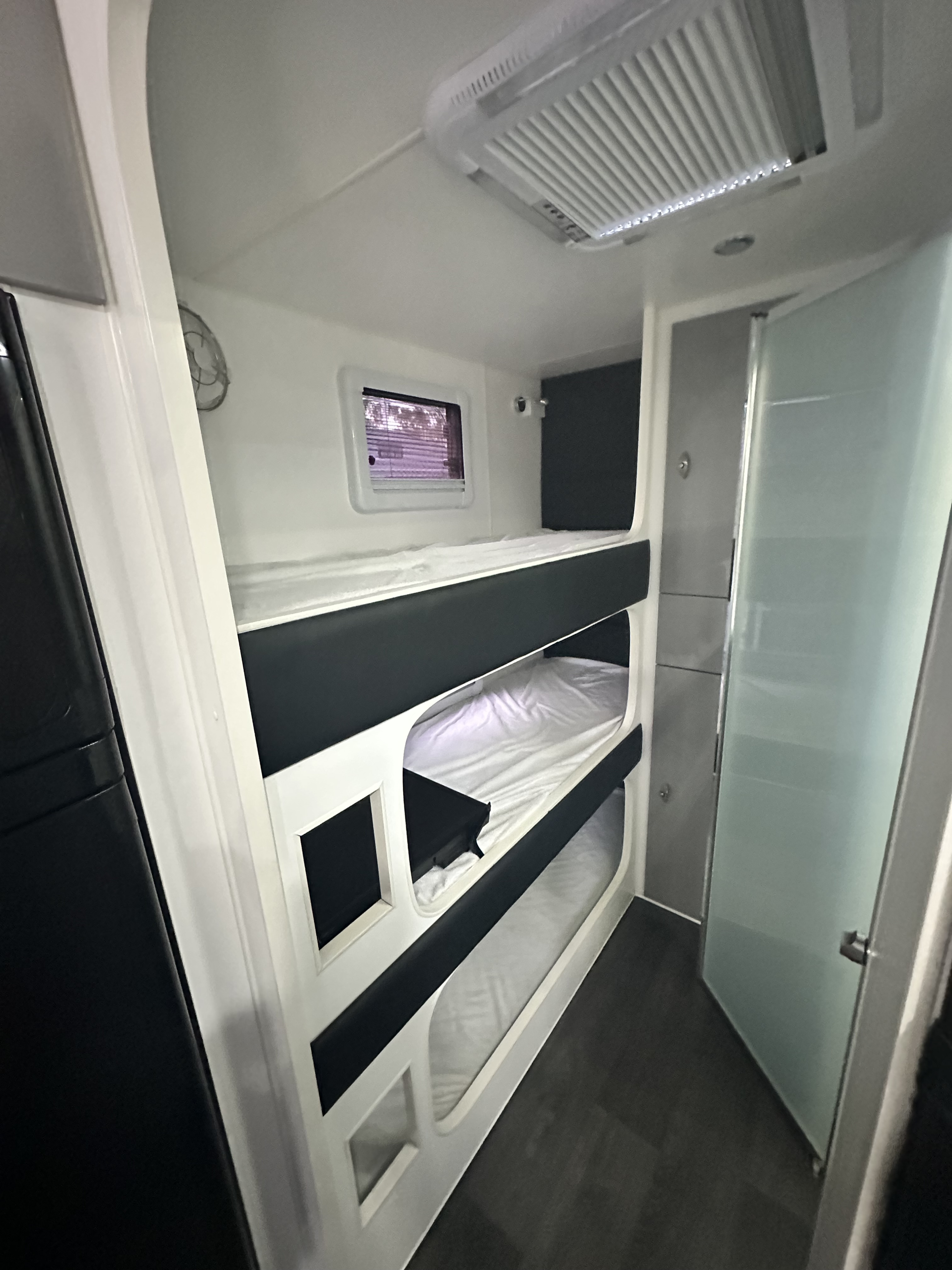 Inside this modern RV rental: a cozy triple bunk setup, overhead A/C, and a frosted glass door leading to a bathroom.. Prime RV Family Base Camp 2023