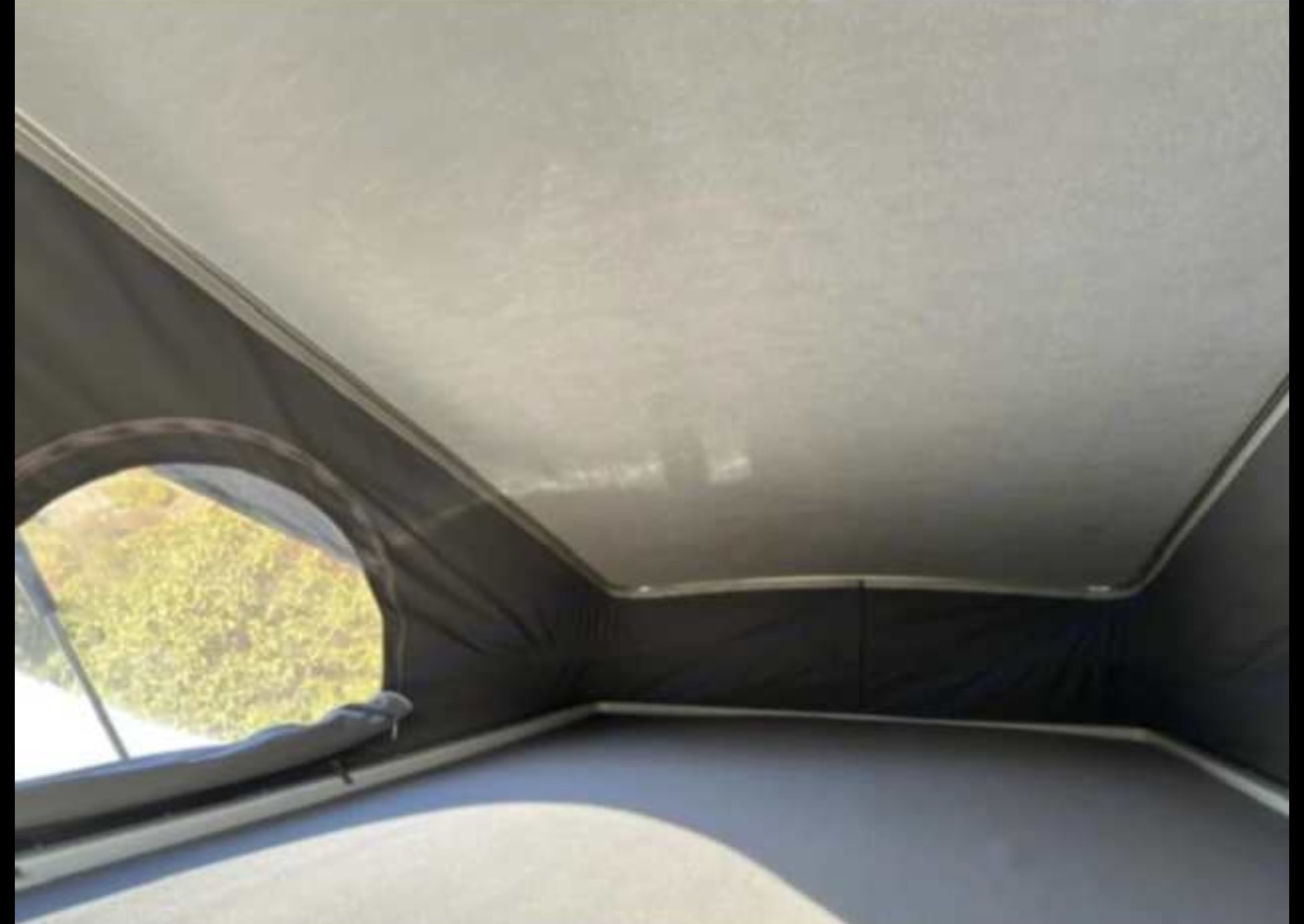 Interior of an RV rental pop-top sleeping tent. Features a light ceiling, dark fabric walls, and a round window showing outside greenery.. Winnebago Solis 2022