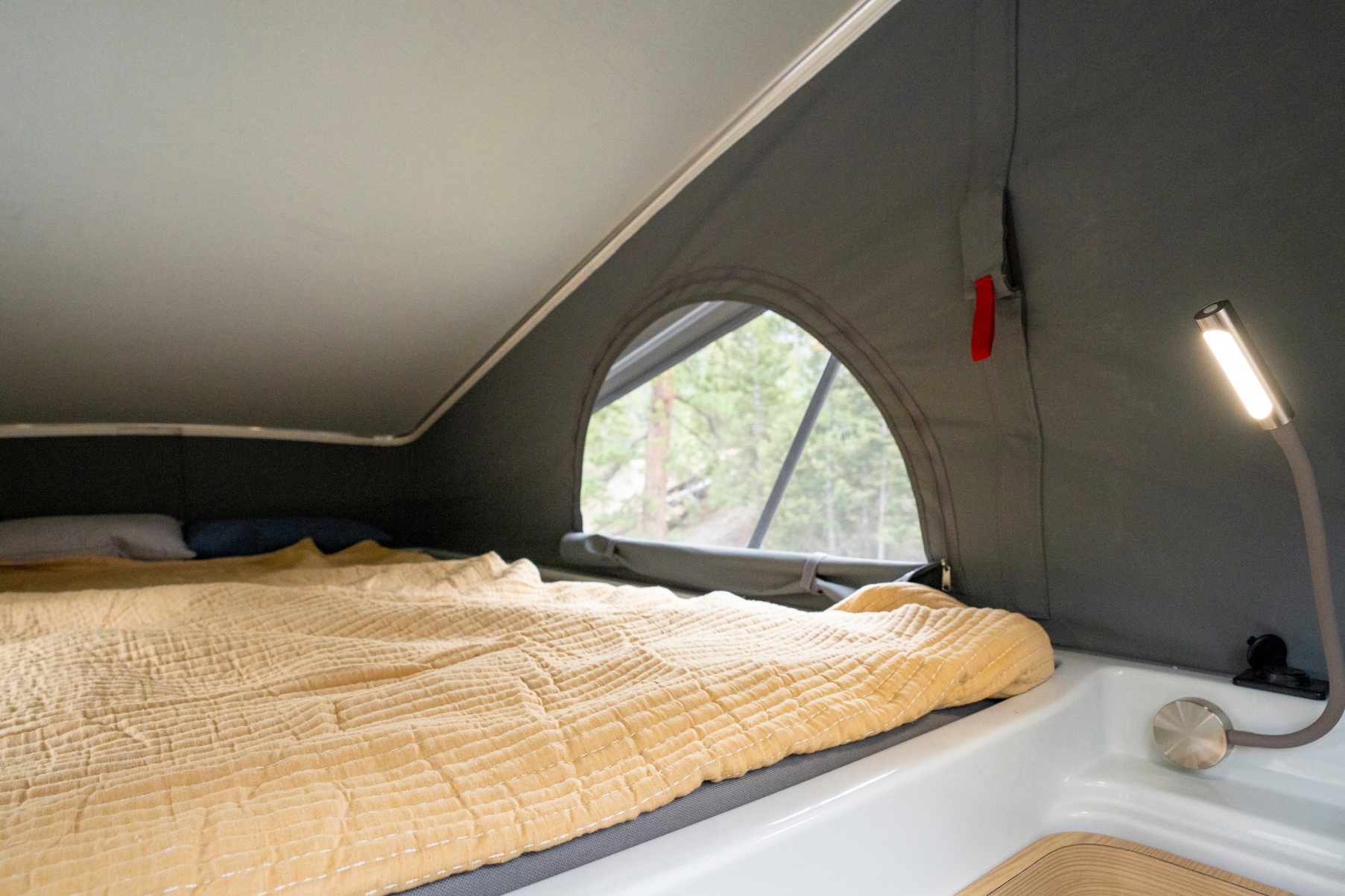 Pop-top RV rental's cozy sleeping area: bed with yellow blanket, forest window view, and reading lamp.. Noovo Pop 2025