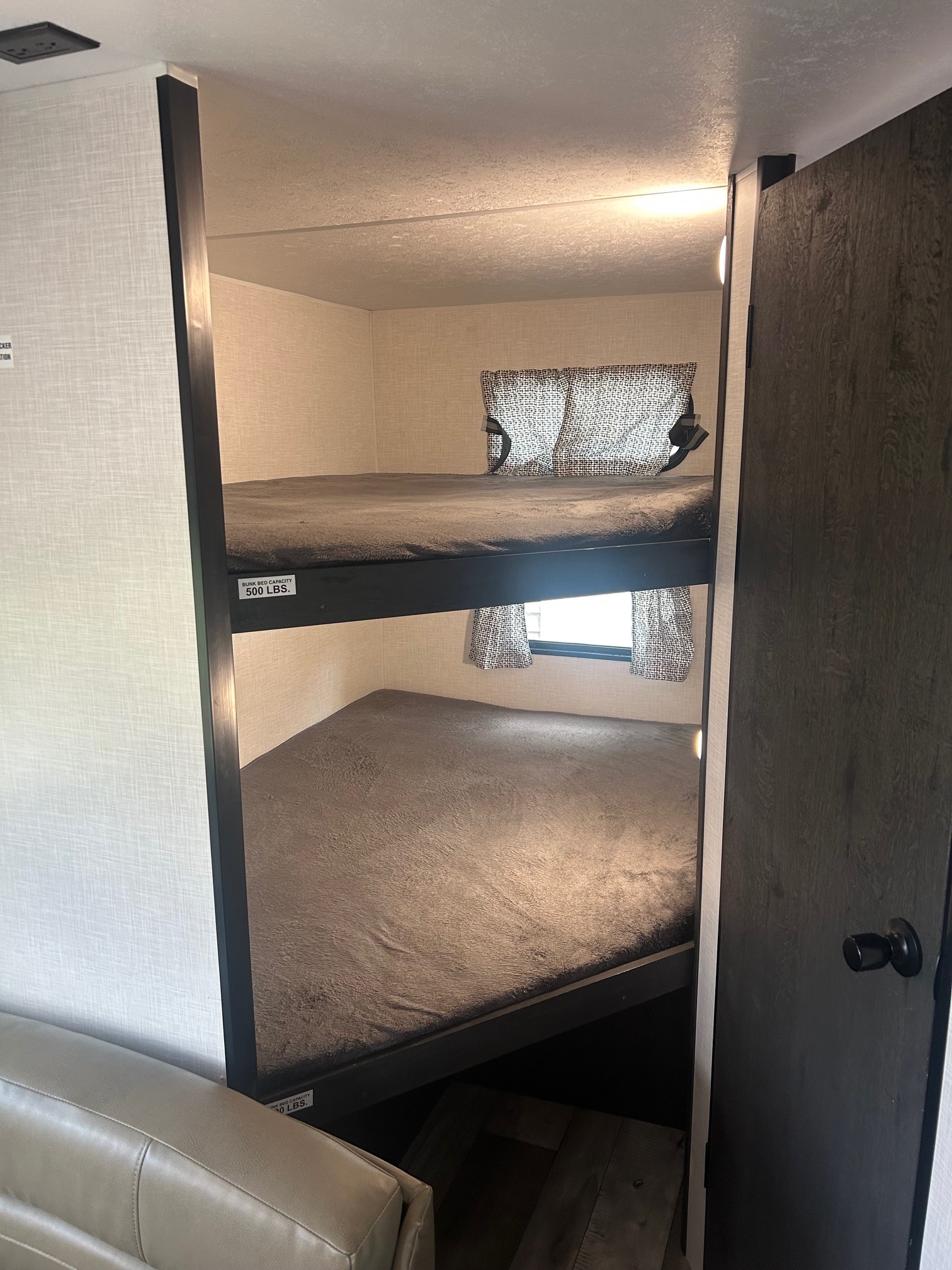 Two-tier bunk beds with soft bedding and small windows inside an RV rental, featuring a 500 lbs capacity label on the top bunk.. Sportsmen SE 2023