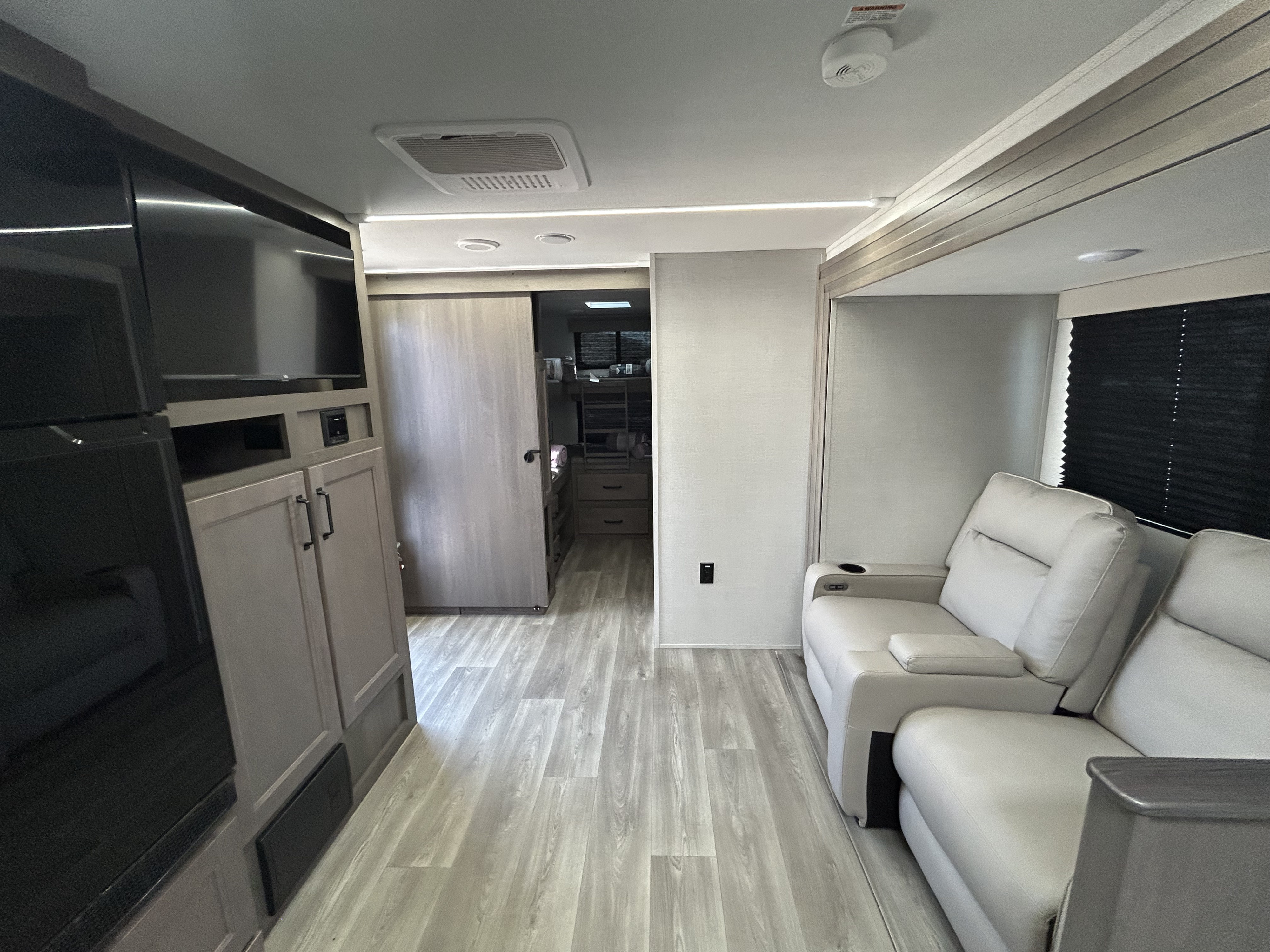 This RV rental interior features light wood-look flooring, cream recliners, a TV, fridge, and a hallway to a rear room.. Grand Design Transcend Xplor 2024