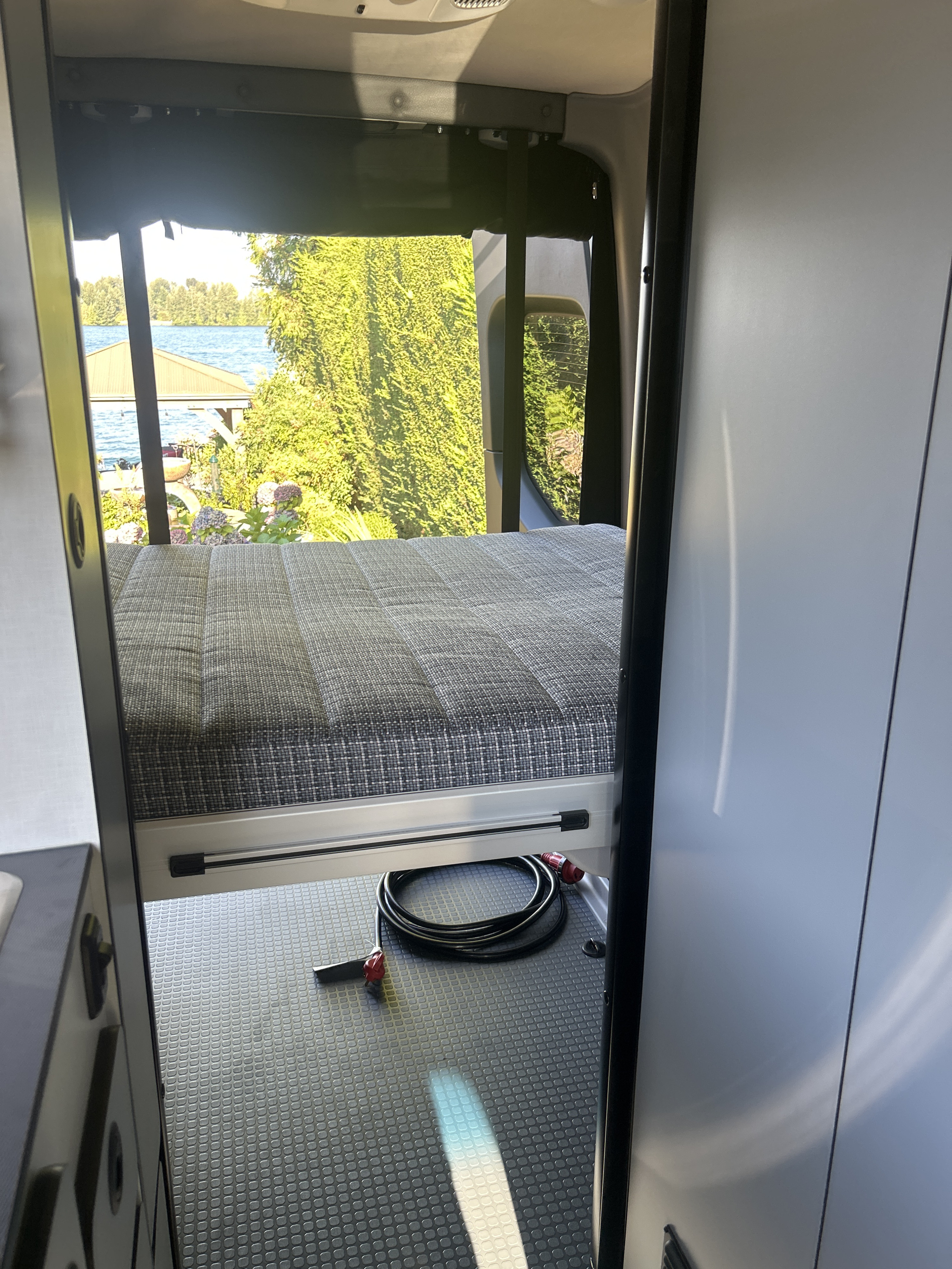 Enjoy an RV rental! Camper van interior with comfy bed, scenic lake view, and shore power cord on patterned floor.. Winnebago Revel 2023