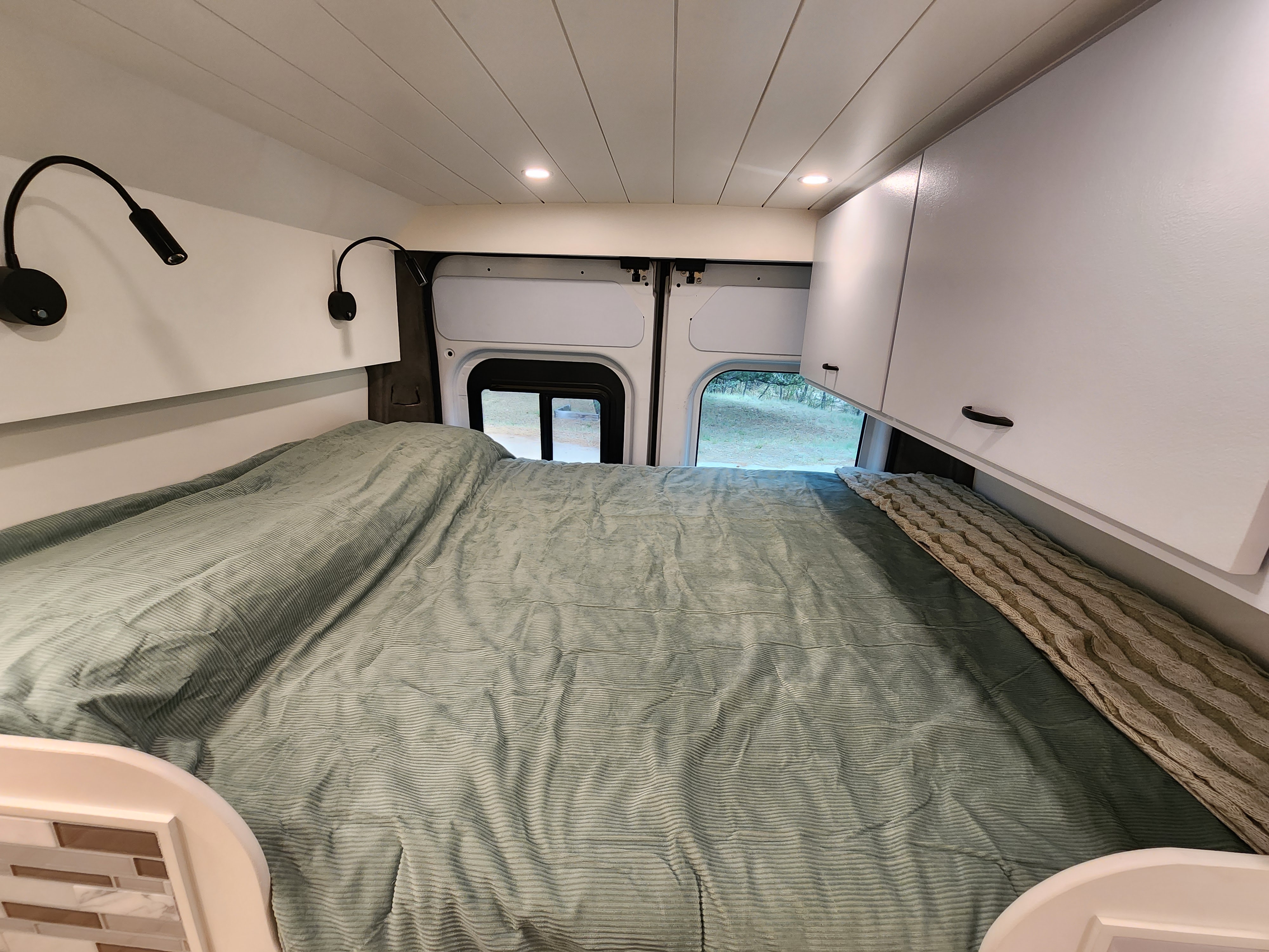 Inside an RV rental, a large bed with a green comforter is framed by reading lights and overhead cabinets. A window reveals the outdoors.. RAM Promaster 2022