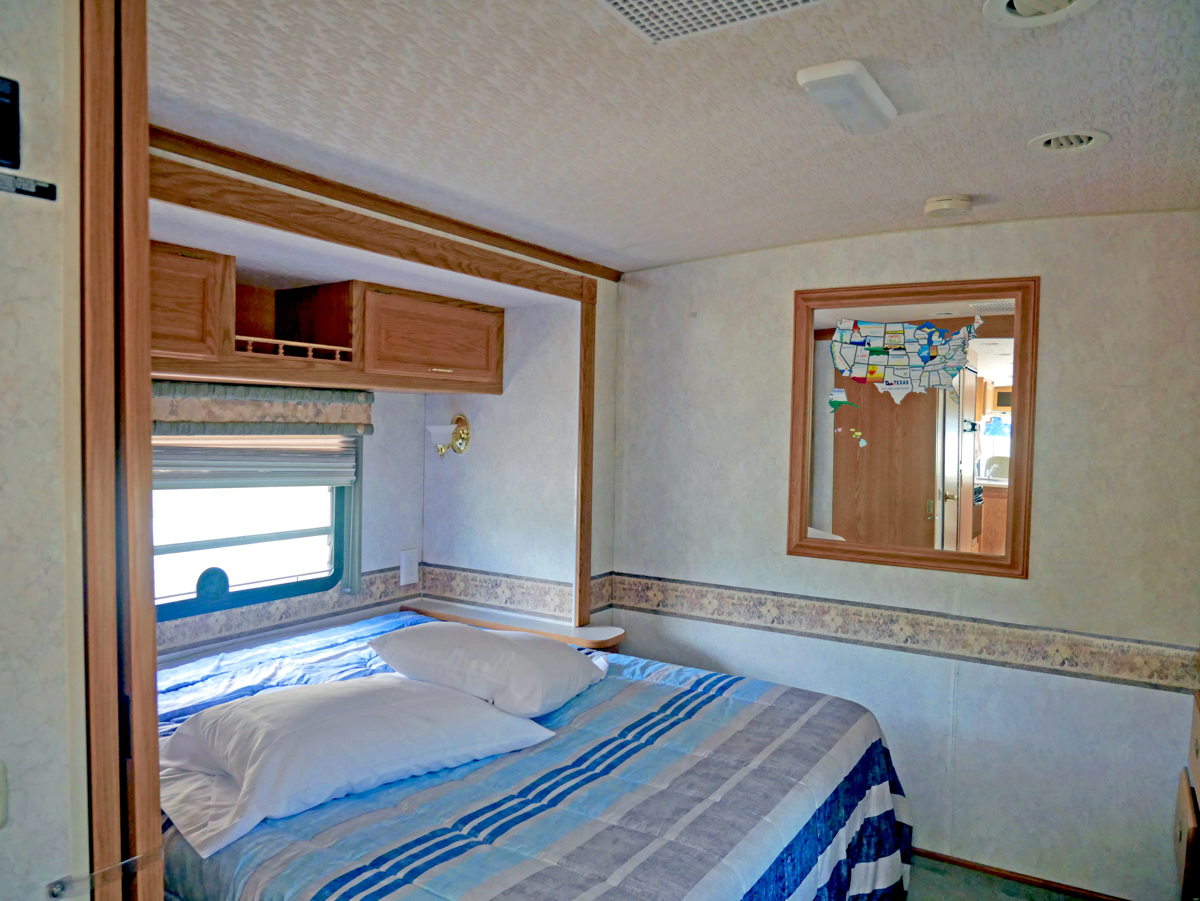 Spacious RV rental bedroom featuring a comfy bed with blue bedding, window, wood cabinets, and a mirror with a US map.. Winnebago Brave 2002