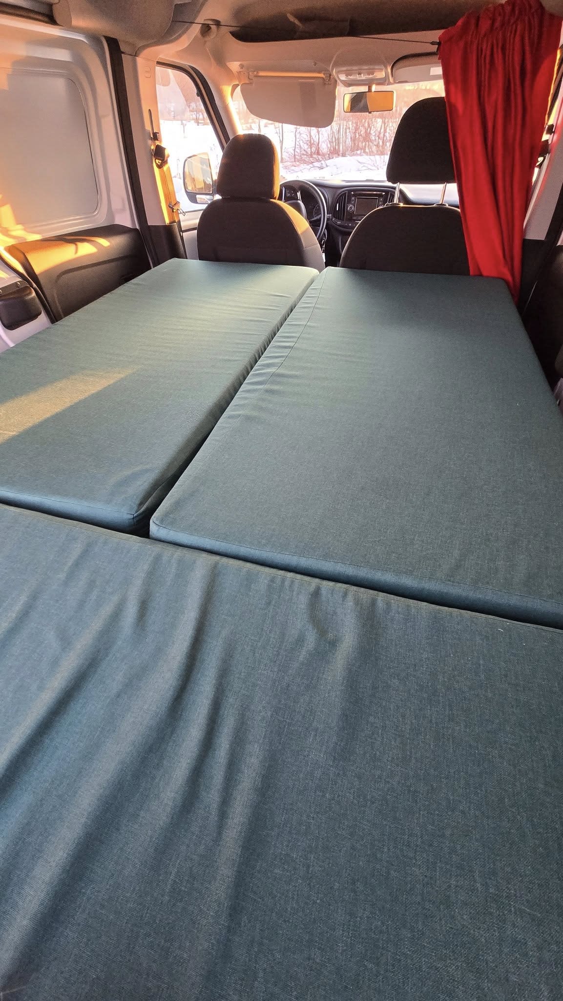 Inside an RV rental: Cozy van bed setup with green cushions for car camping. Snowy view.. Dodge Promaster City 2021