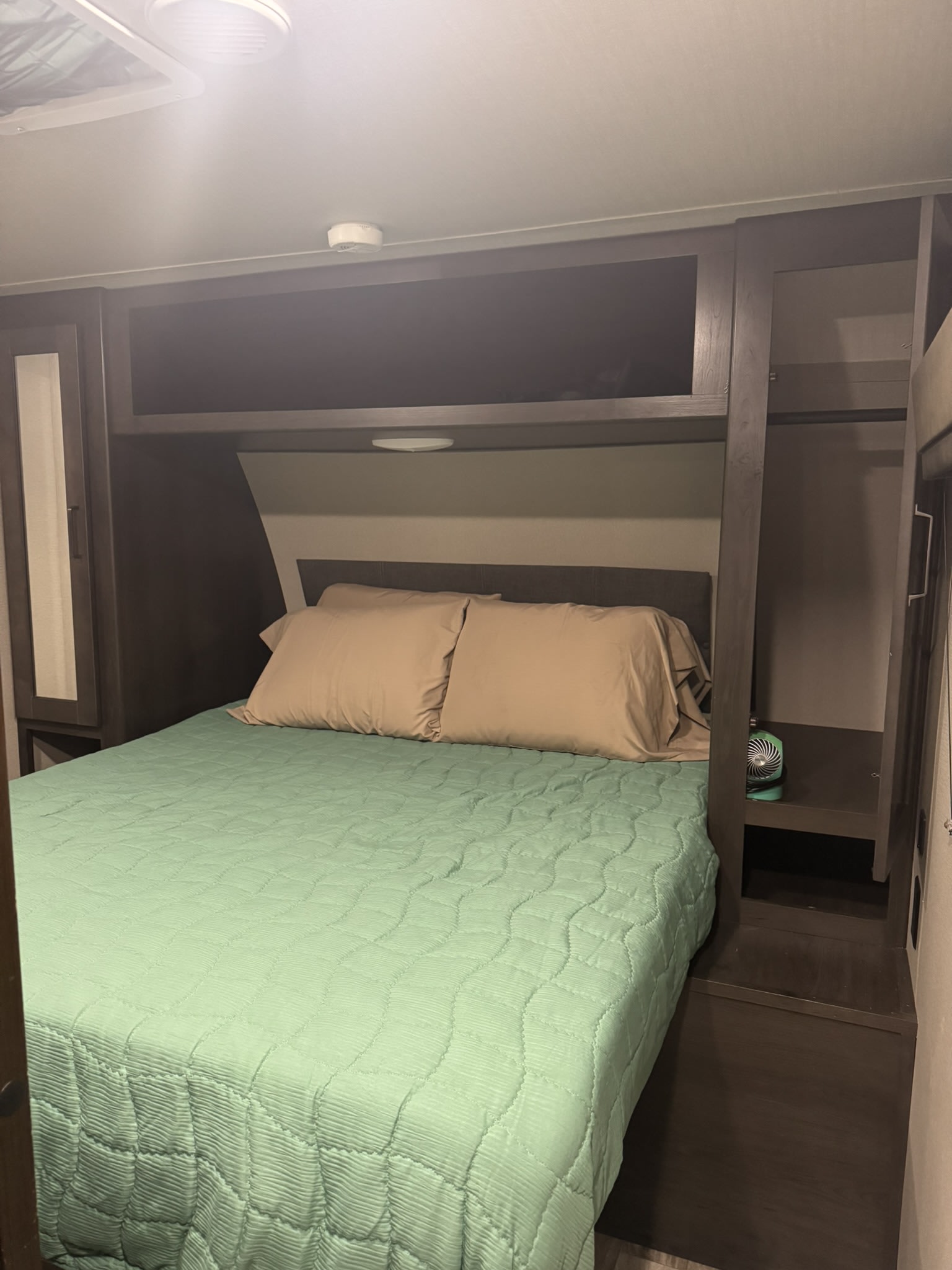 This RV rental bedroom has a green quilted bed, tan pillows, and dark wood built-in storage, plus a ceiling smoke detector.. Grand Design Transcend Xplor 2021