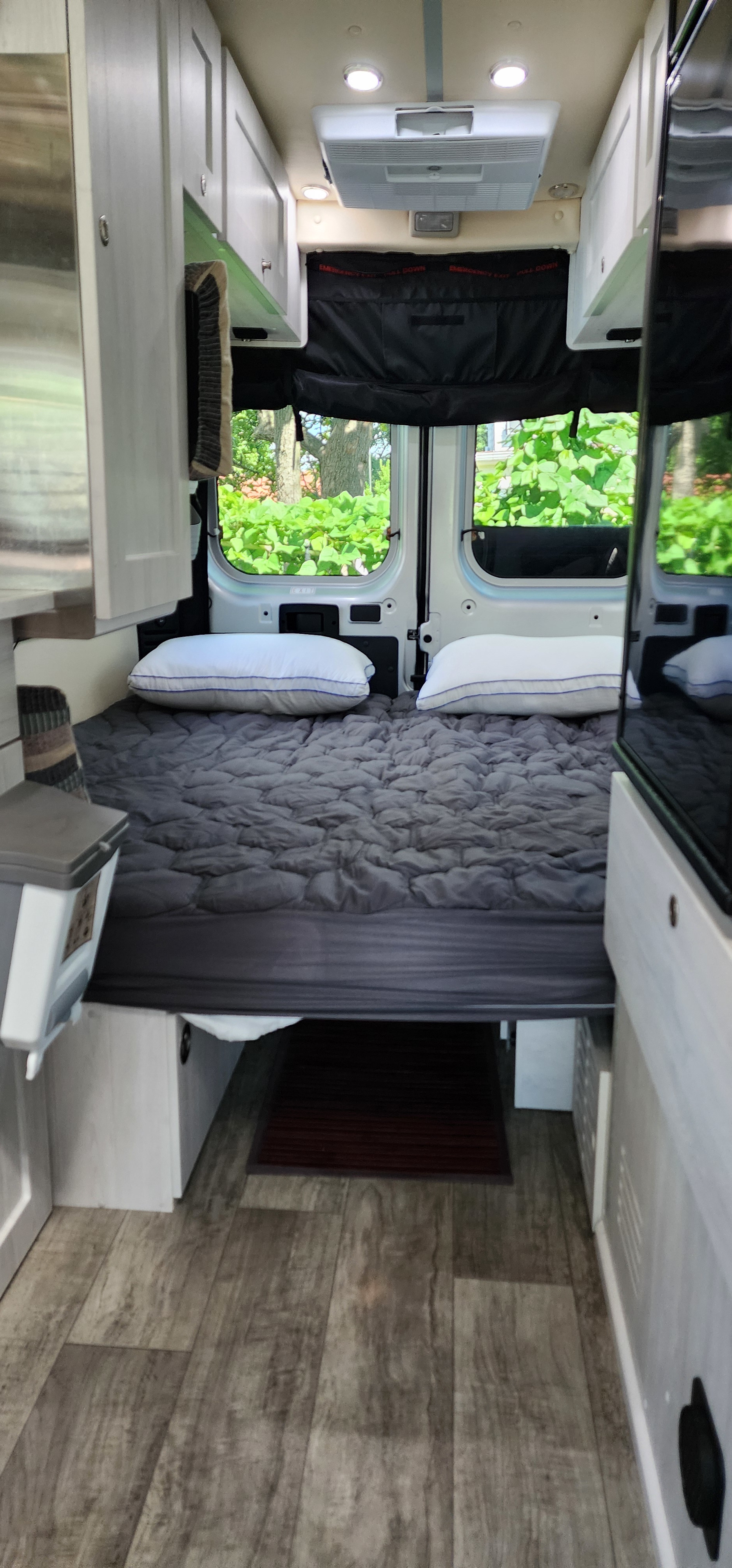 RV rental camper van interior: cozy bed with grey comforter, overhead storage, AC, and rear windows showing greenery.. Roadtrek Roadtrek Class B 2023