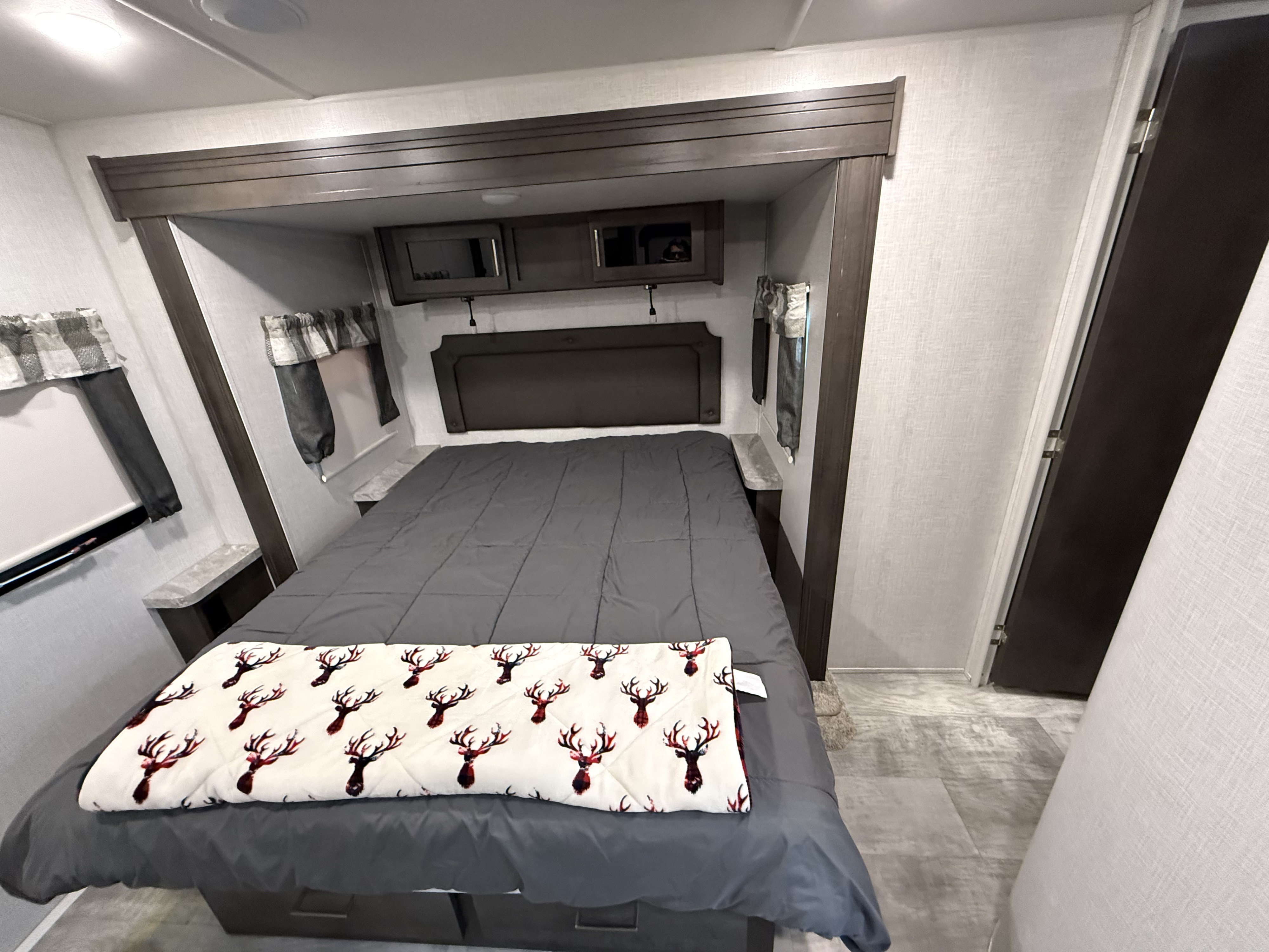 Cozy RV rental bedroom with a grey bed, deer-patterned blanket, overhead cabinets, and built-in side shelves.. Forest River Flagstaff Super Lite 2021