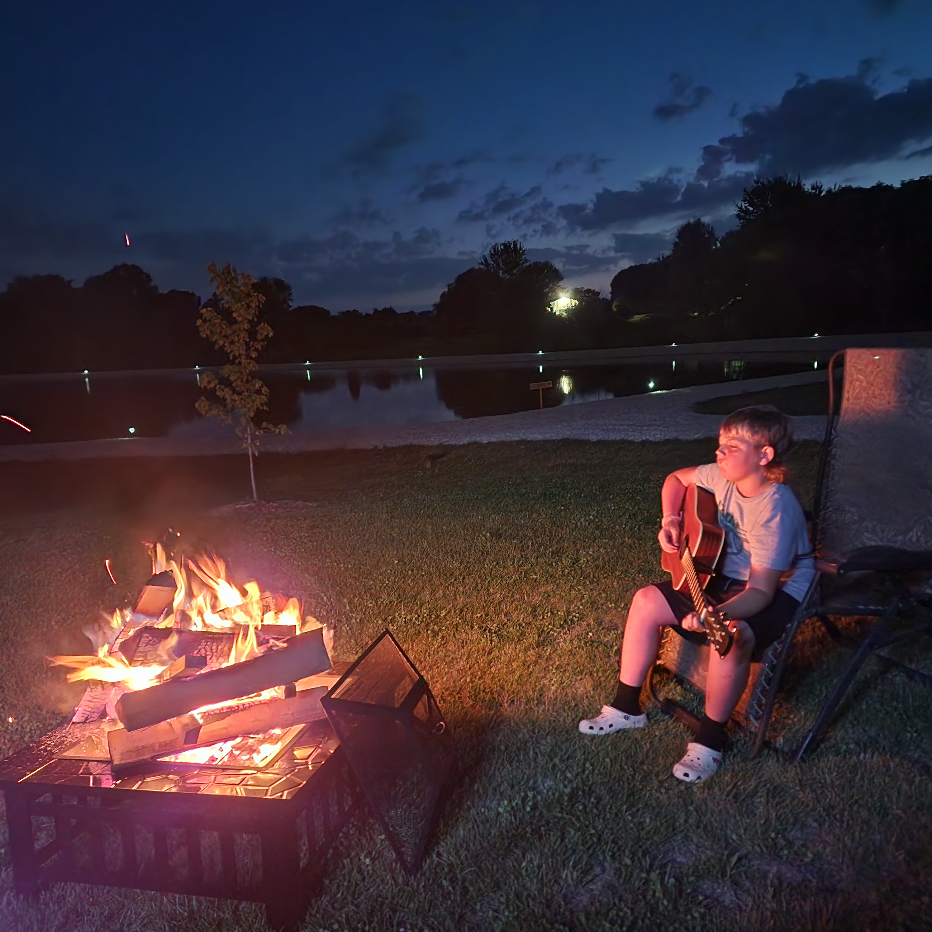 Serene evening at a lake: a young person plays guitar by a crackling bonfire, ideal after an RV rental trip.. Keystone Passport 2018
