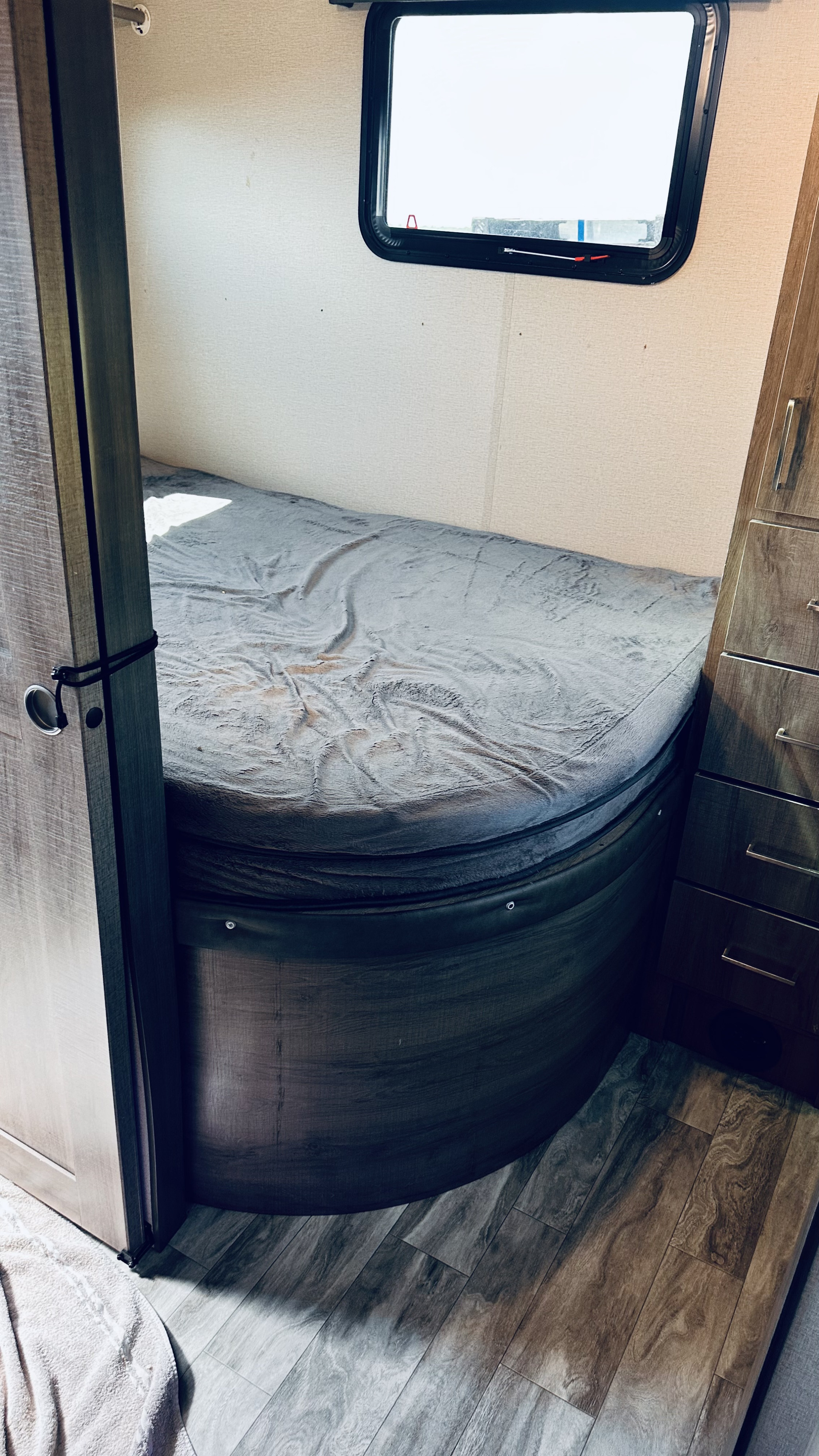 Inside an RV rental bedroom: a cozy bed with gray blanket, window, and wooden storage cabinet.. Grand Design Imagine 2021