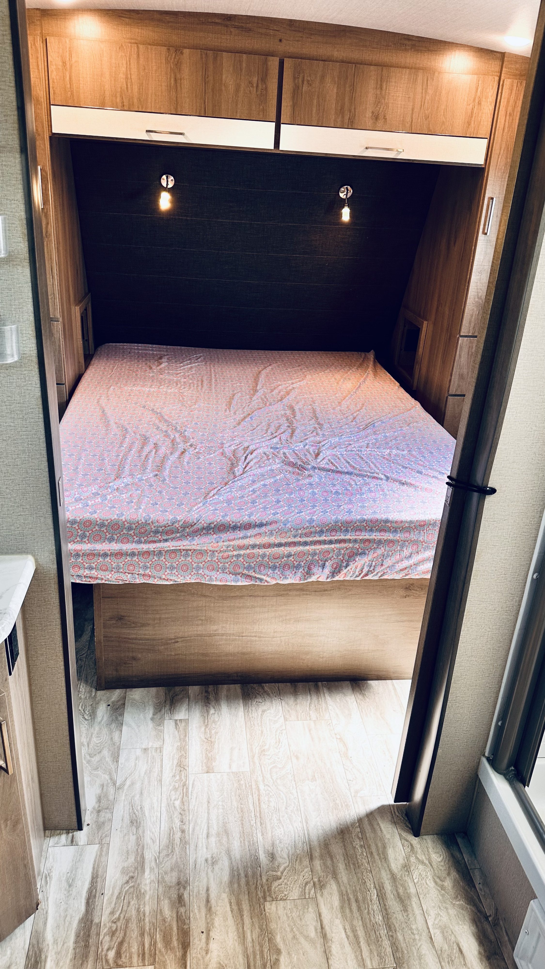 View inside an RV rental bedroom showing a bed with patterned sheets, overhead cabinets, and reading lights.. Grand Design Imagine 2021